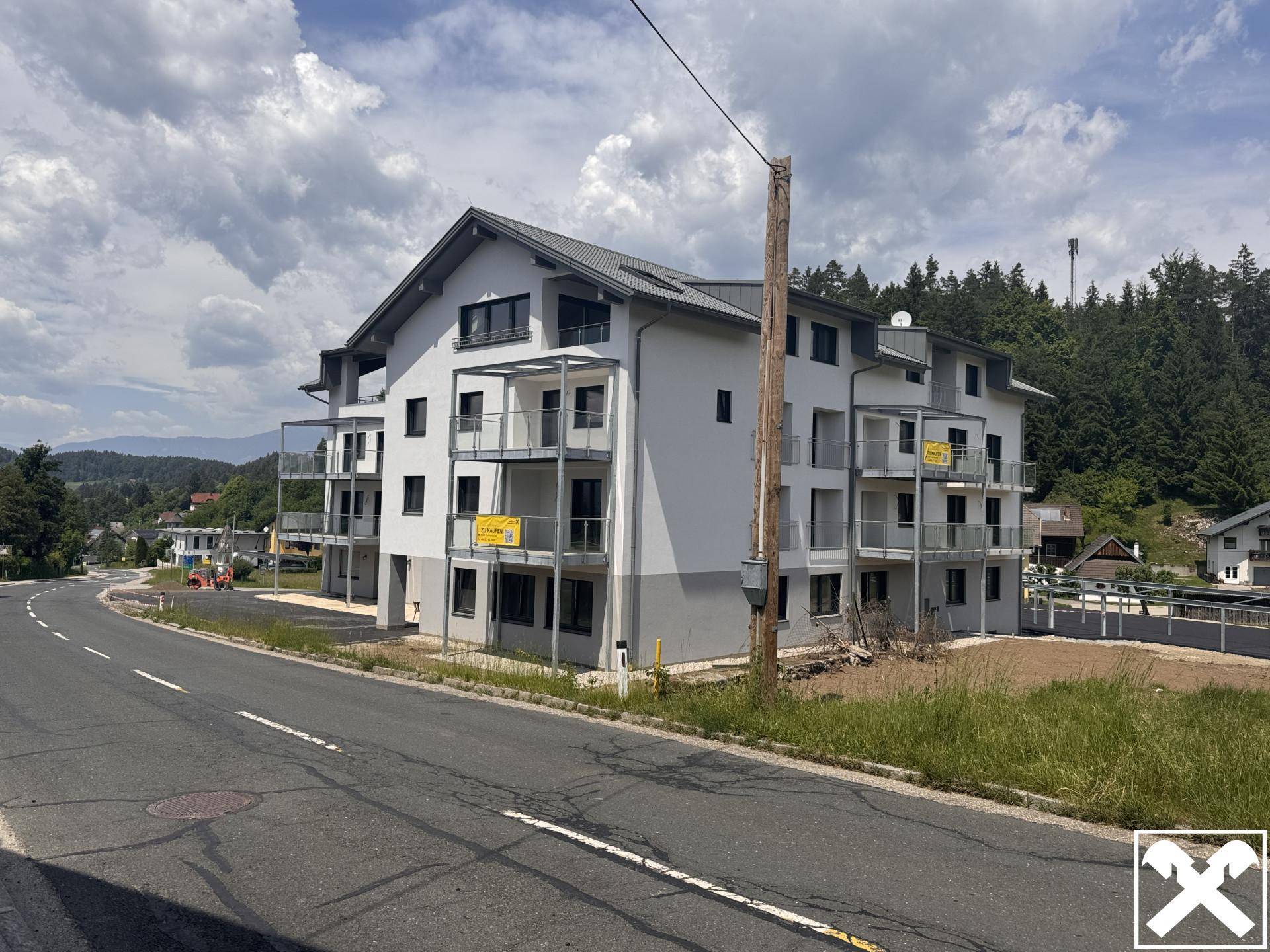 Neubau-Wohngebäude an einer Straße, umgeben von grüner Landschaft und bewölktem Himmel.