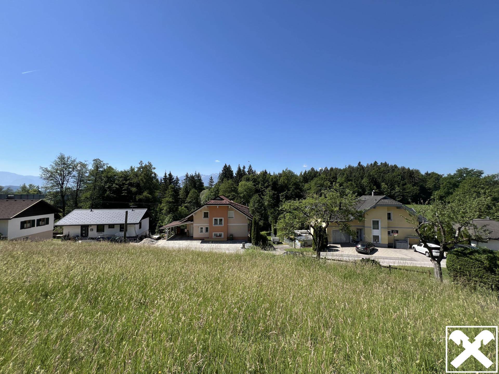 Panoramablick über eine grüne Wiese auf mehrere Wohnhäuser und bewaldete Hügel.