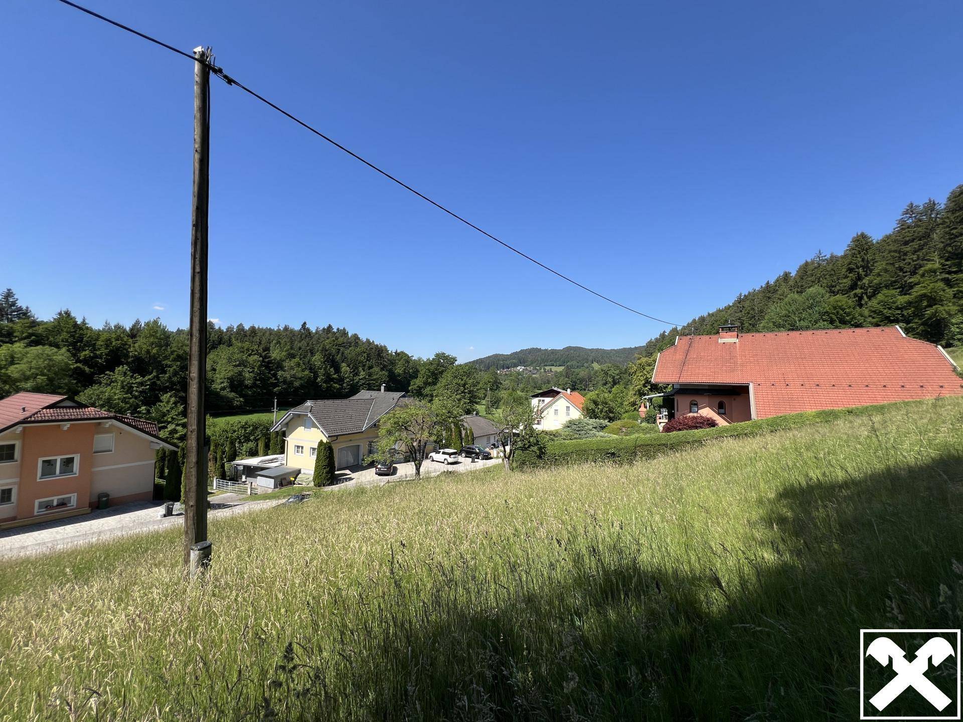 Weitläufige grüne Wiese mit Blick auf Wohnhäuser und bewaldete Hügel in der Ferne.