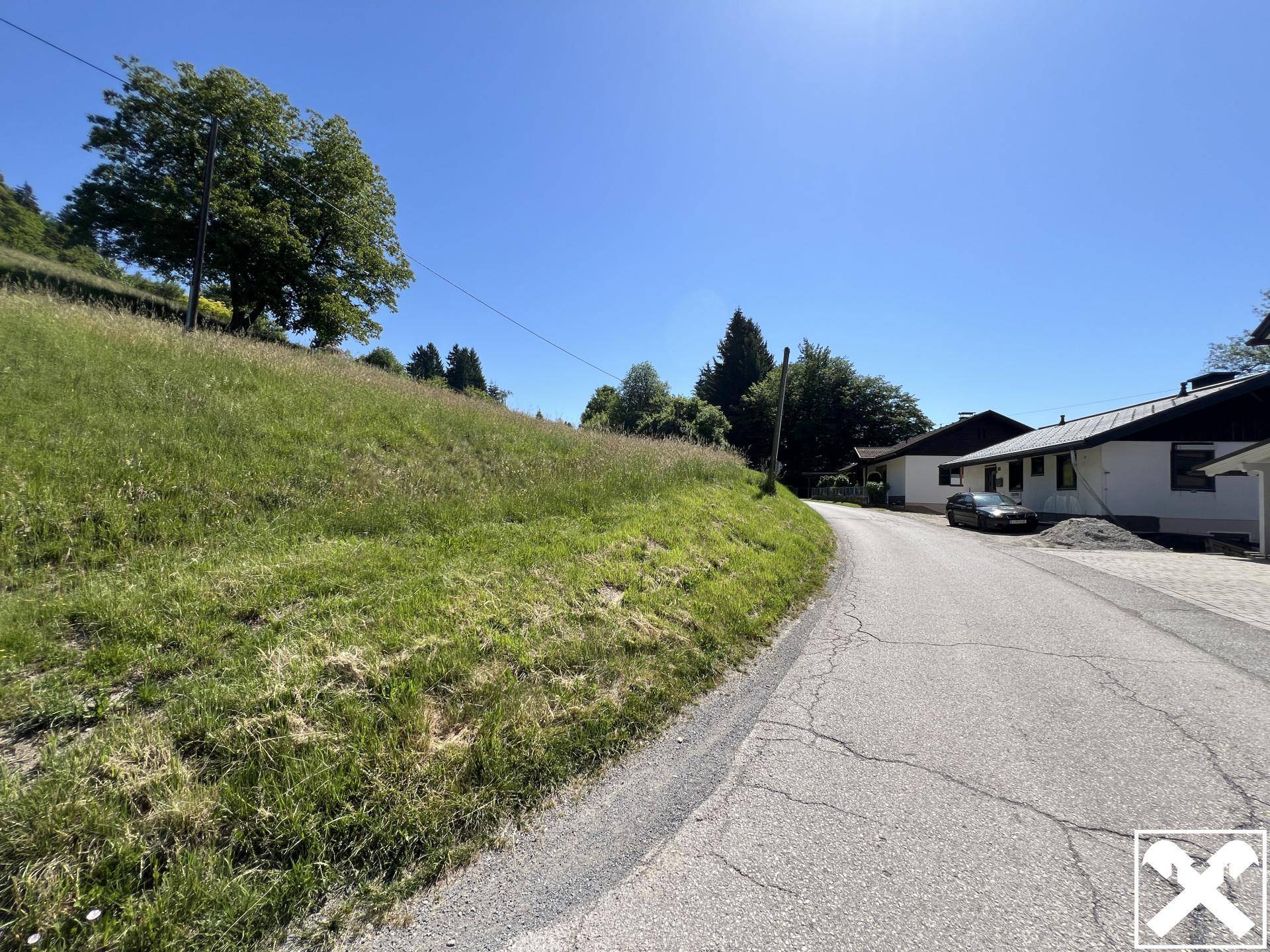 Grüne Böschung neben einer asphaltierten Straße mit Häusern und Bäumen im Hintergrund.