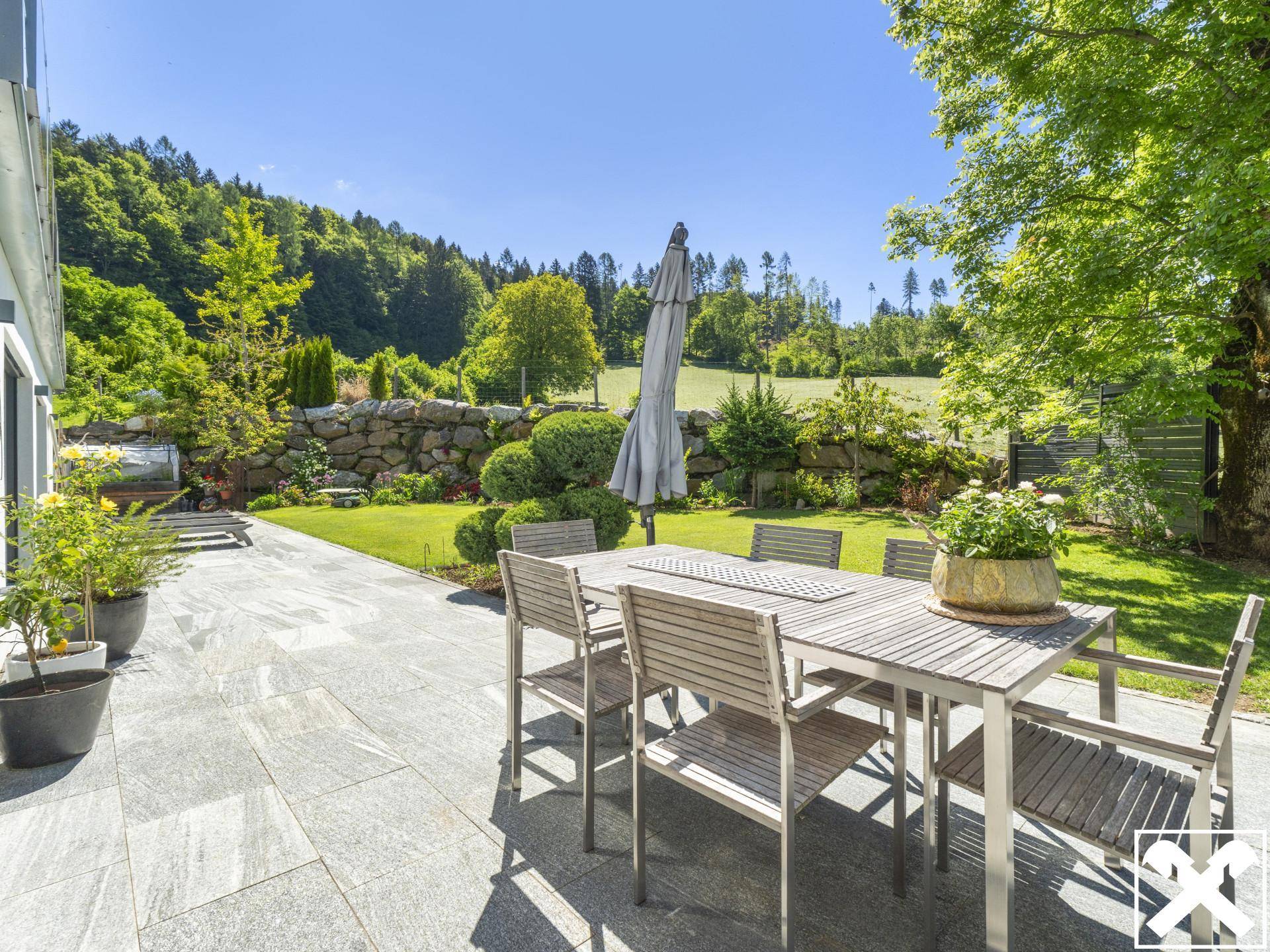 Geräumige Terrasse mit Esstisch und Stühlen, umgeben von üppigem Grün und einer Steinmauer.