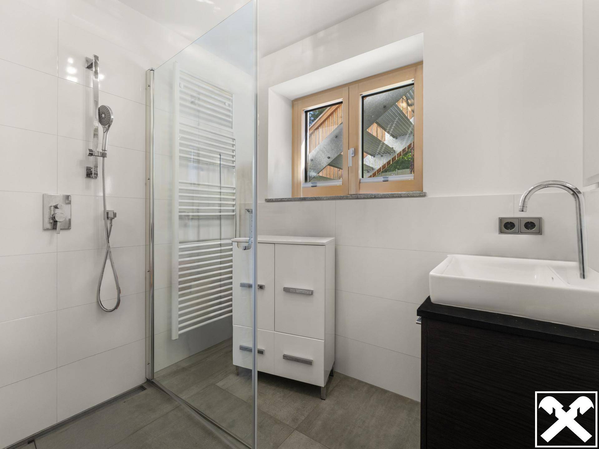 Helles Badezimmer mit Dusche, Waschtisch und Fenster, das viel Tageslicht hereinlässt.