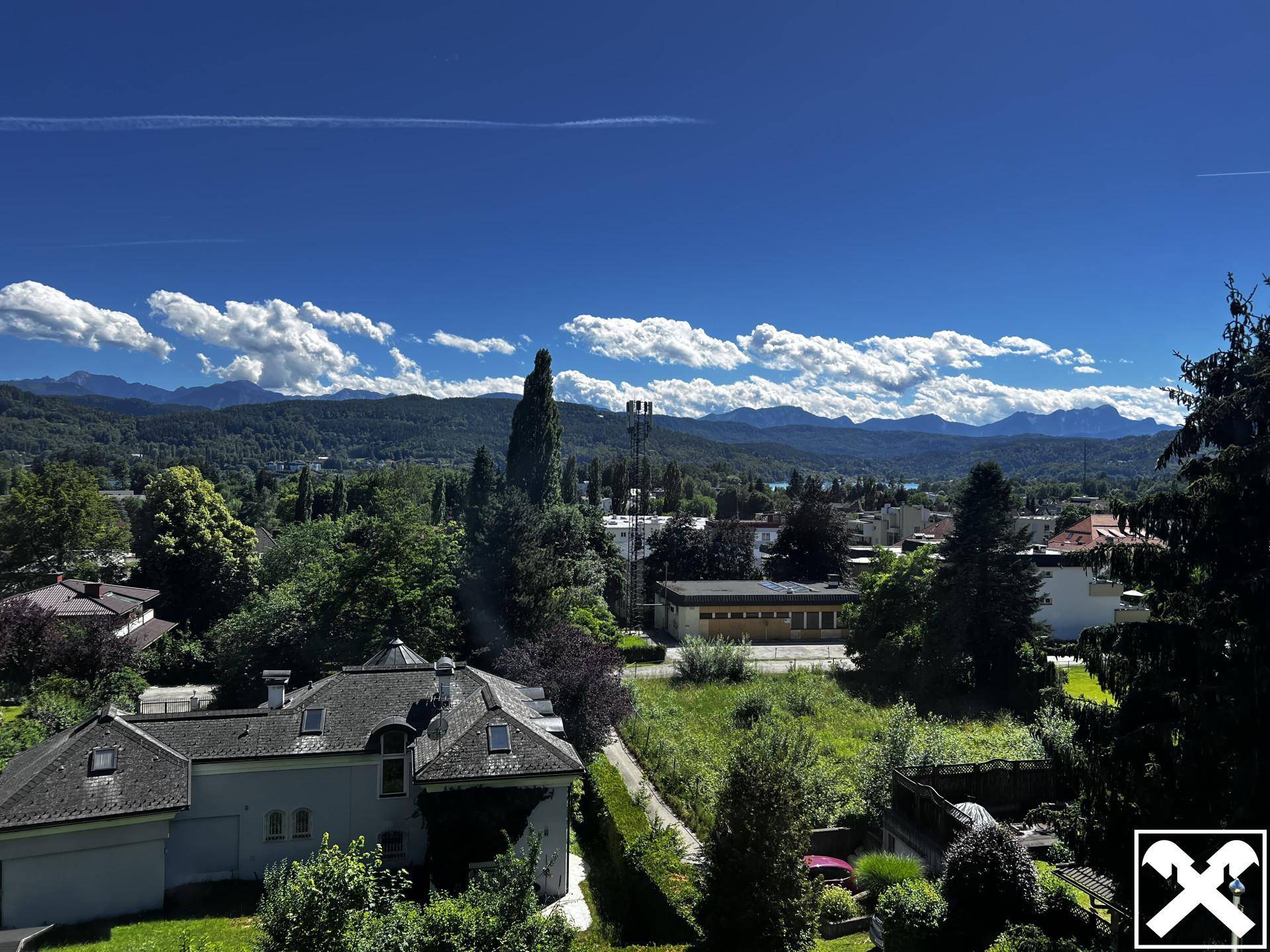 Panoramablick über die grüne Landschaft, Dächer und die umliegenden Berge unter blauem Himmel.