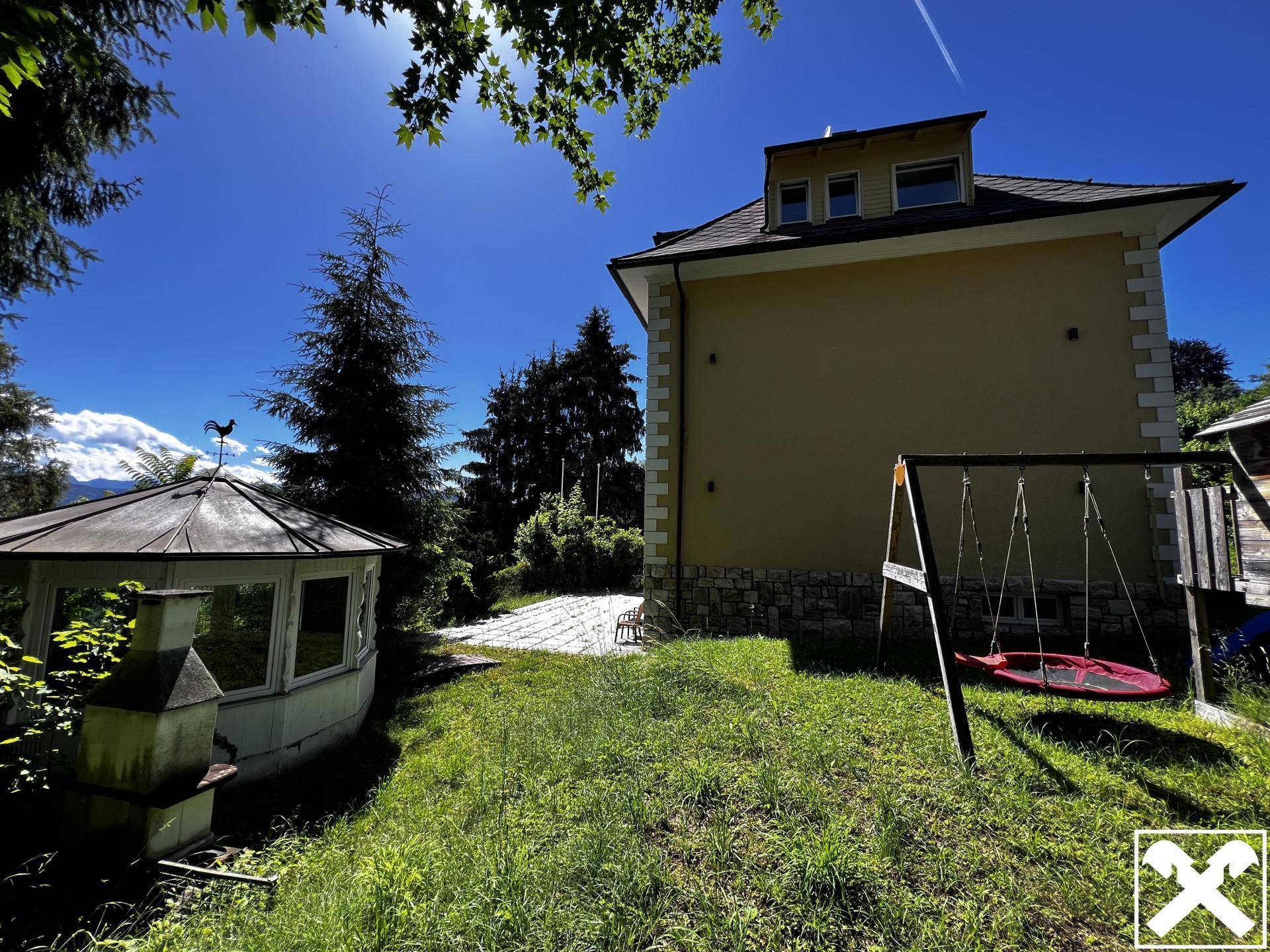 Seitlicher Blick auf das Haus mit Garten, Pavillon, Grillplatz und Kinderschaukel unter blauem Himmel.