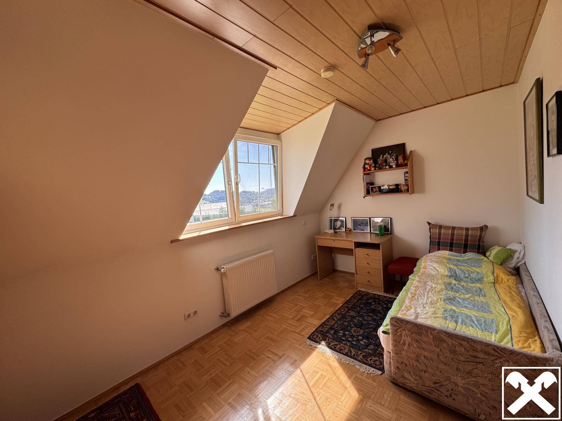 Helles Schlafzimmer im Dachgeschoss mit Holzboden, Schreibtisch und Fenster mit Ausblick.