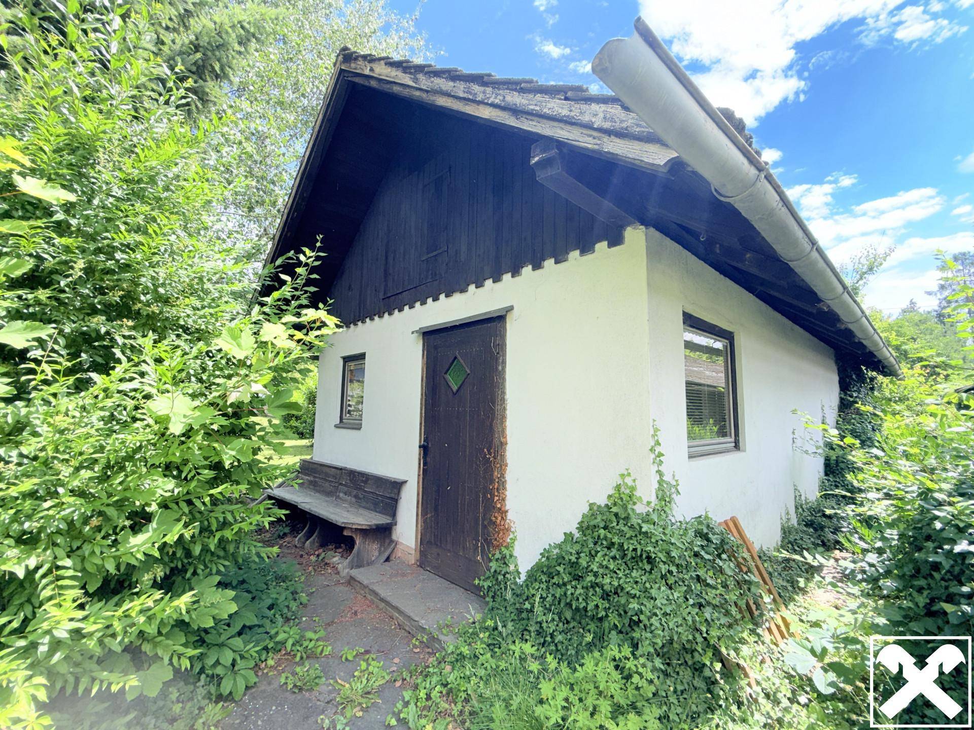 Charmantes, kleines Gartenhaus mit Holztür und Fenster, umgeben von dichtem Bewuchs.