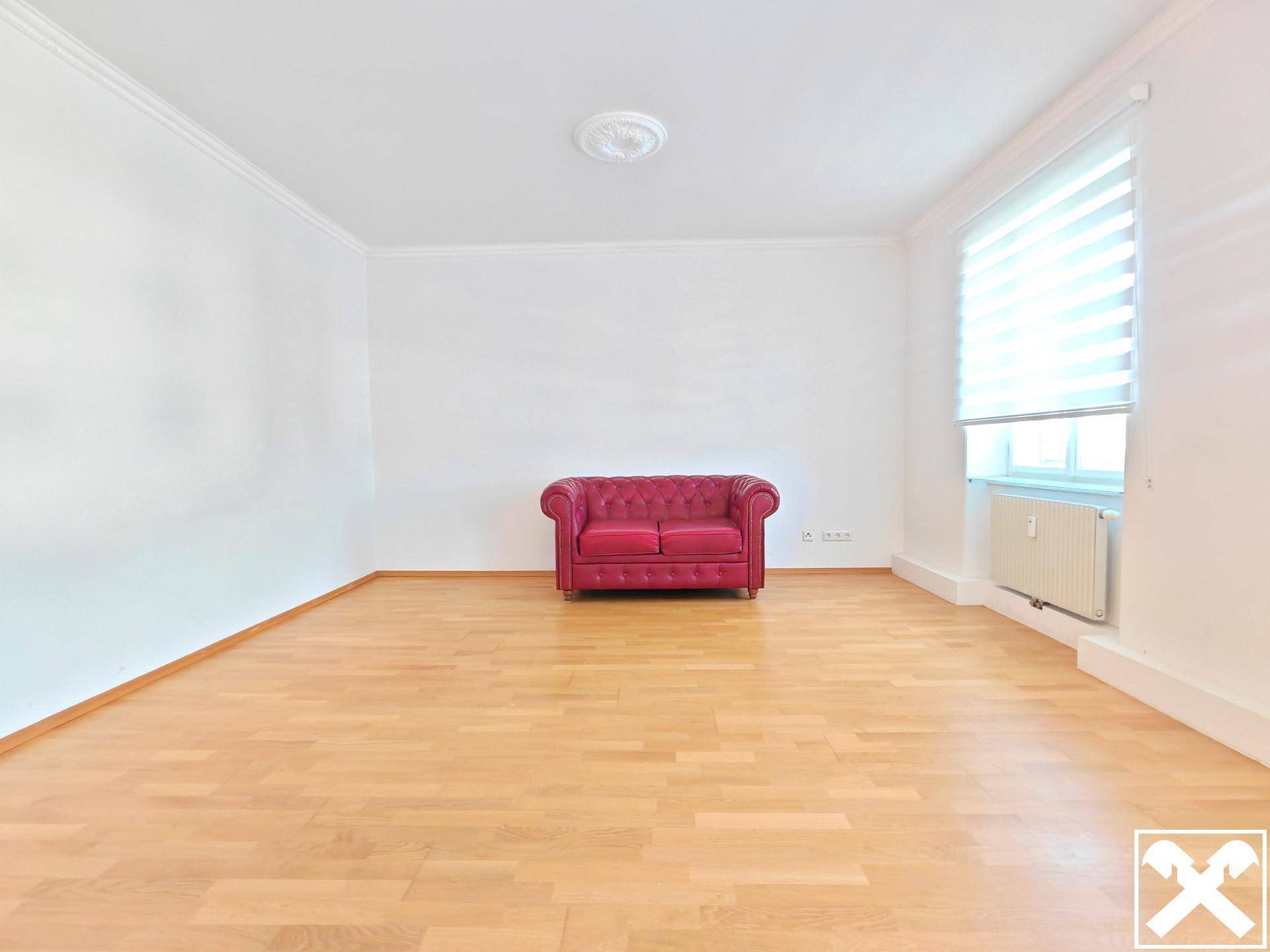 Geräumiges Wohnzimmer mit hellem Parkettboden, weißen Wänden und einem einzelnen roten Sofa.