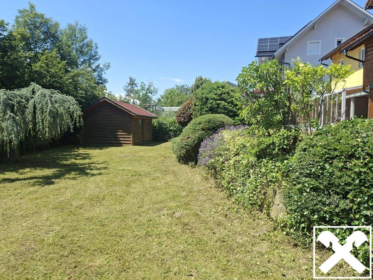 Großer Garten mit Rasenfläche, Sträuchern und einem kleinen Holzschuppen unter blauem Himmel.