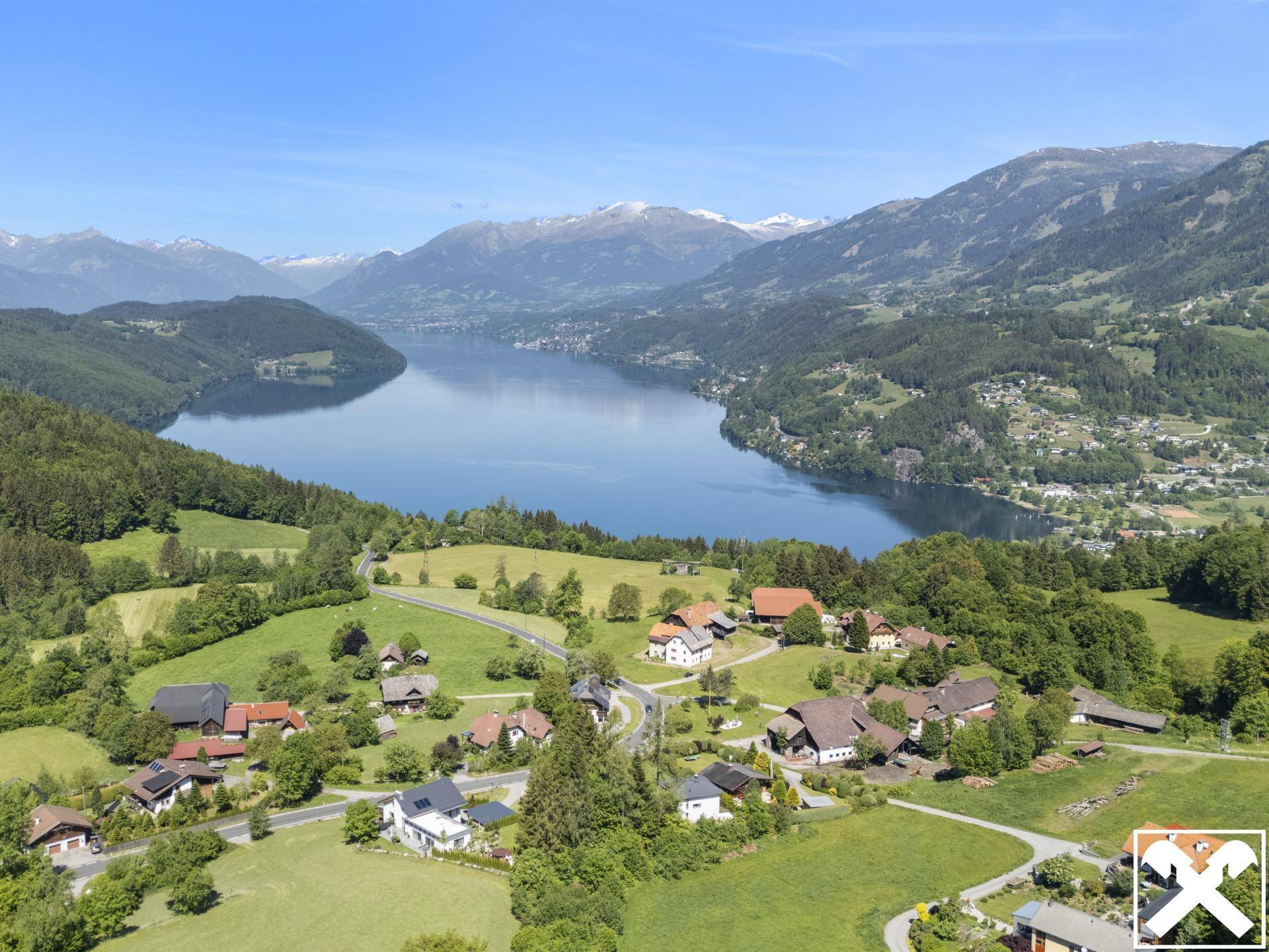 Malerische Luftaufnahme eines Dorfes am Seeufer mit umliegenden Bergen und grünen Hügeln.