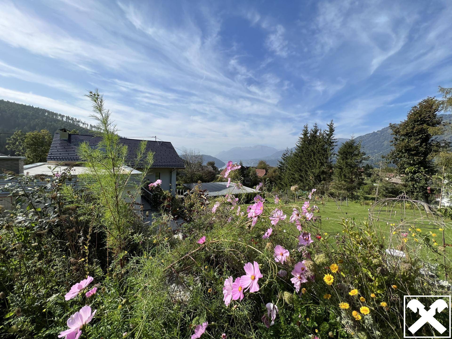 Bunte Blumen im Vordergrund mit Blick auf ein Haus, Berge und den blauen Himmel.