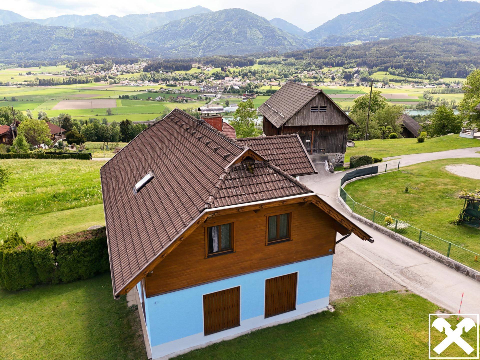 Luftaufnahme des Hauses mit Satteldach und Holzverkleidung, umgeben von grünen Feldern und einem Dorf im Tal.