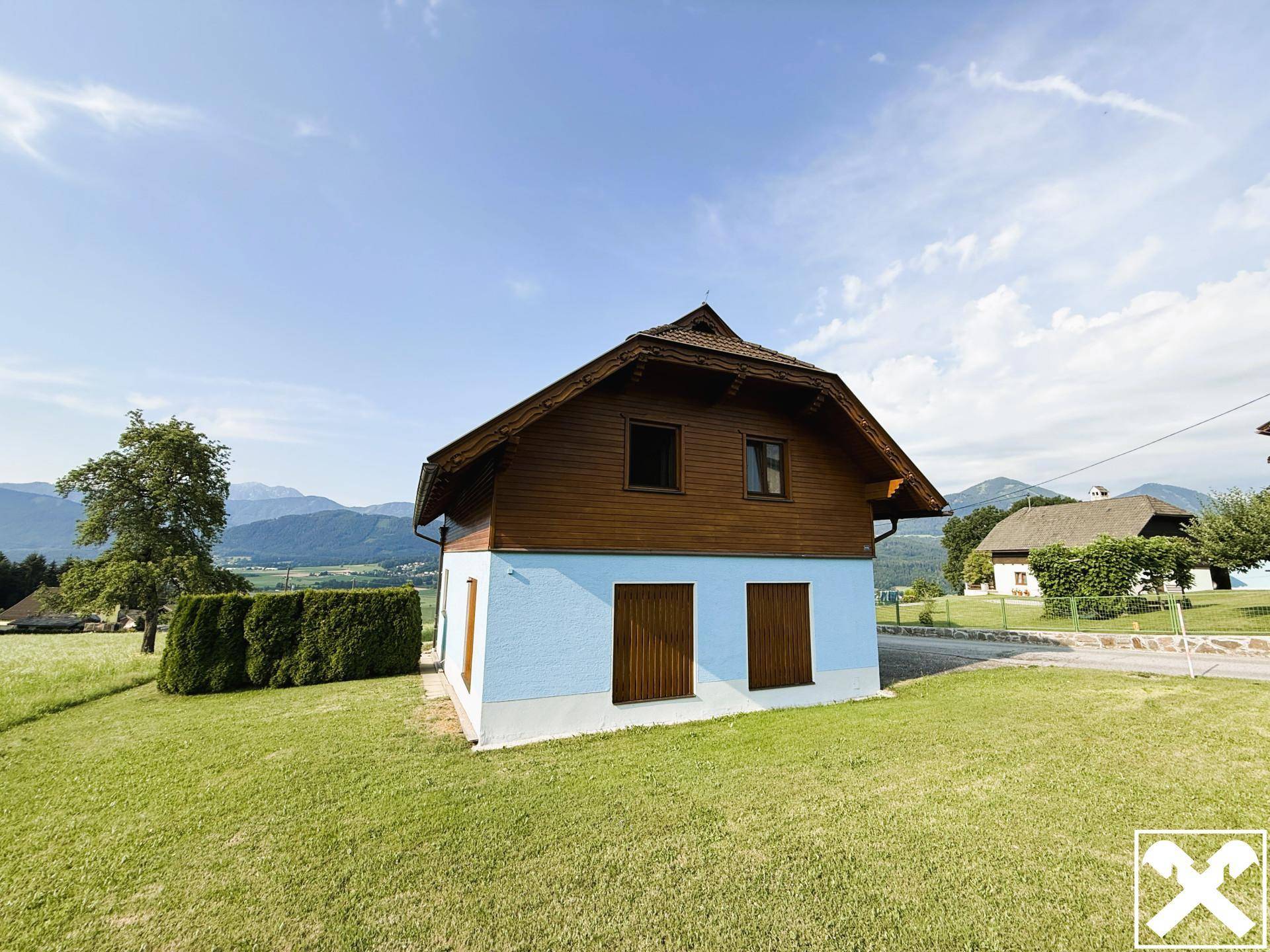 Haus mit blauer Fassade und Holzverkleidung, umgeben von einem gepflegten Rasen und Bergen in der Ferne.