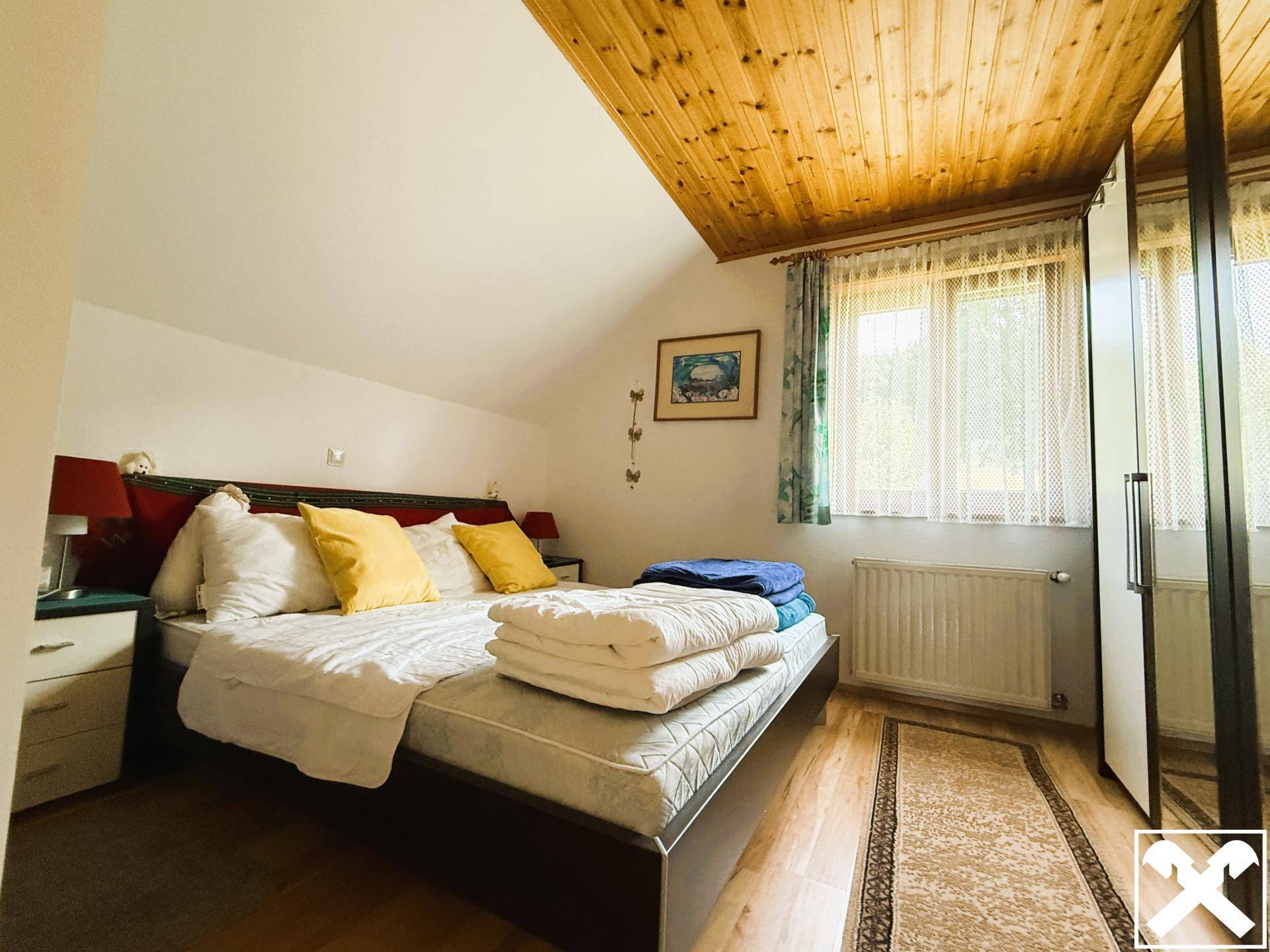 Schlafzimmer mit Doppelbett, Holzdecke und Dachschräge, hell beleuchtet durch ein Fenster.