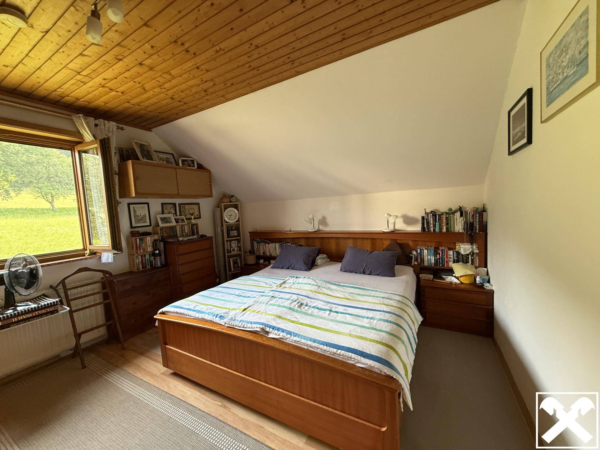Schlafzimmer mit Holzdecke, Doppelbett und Bücherregalen, das durch ein großes Fenster viel Licht erhält.