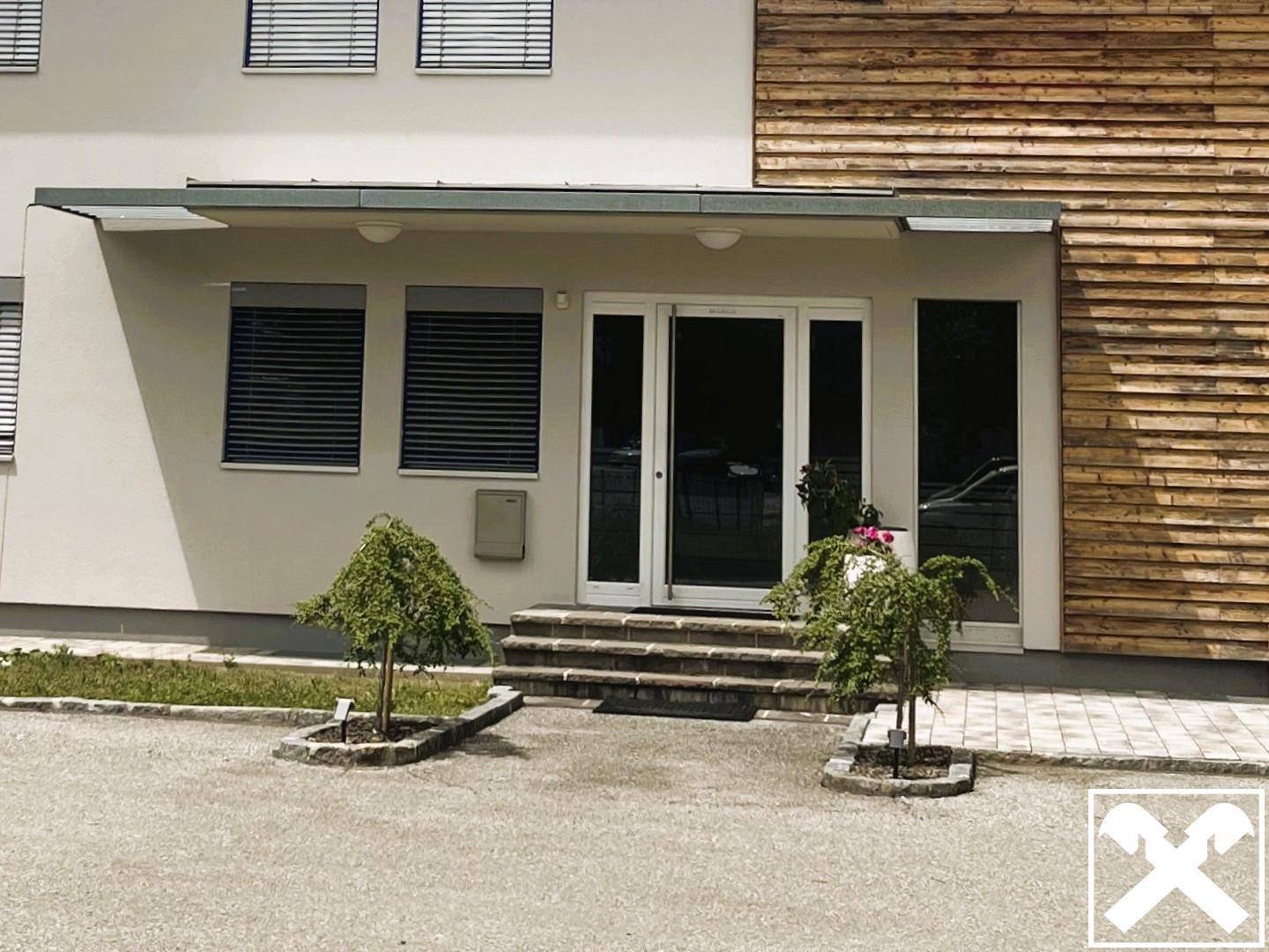 Eingangsbereich des modernen Gebäudes mit Glastüren, Steinstufen und kleinen Zierbäumen.
