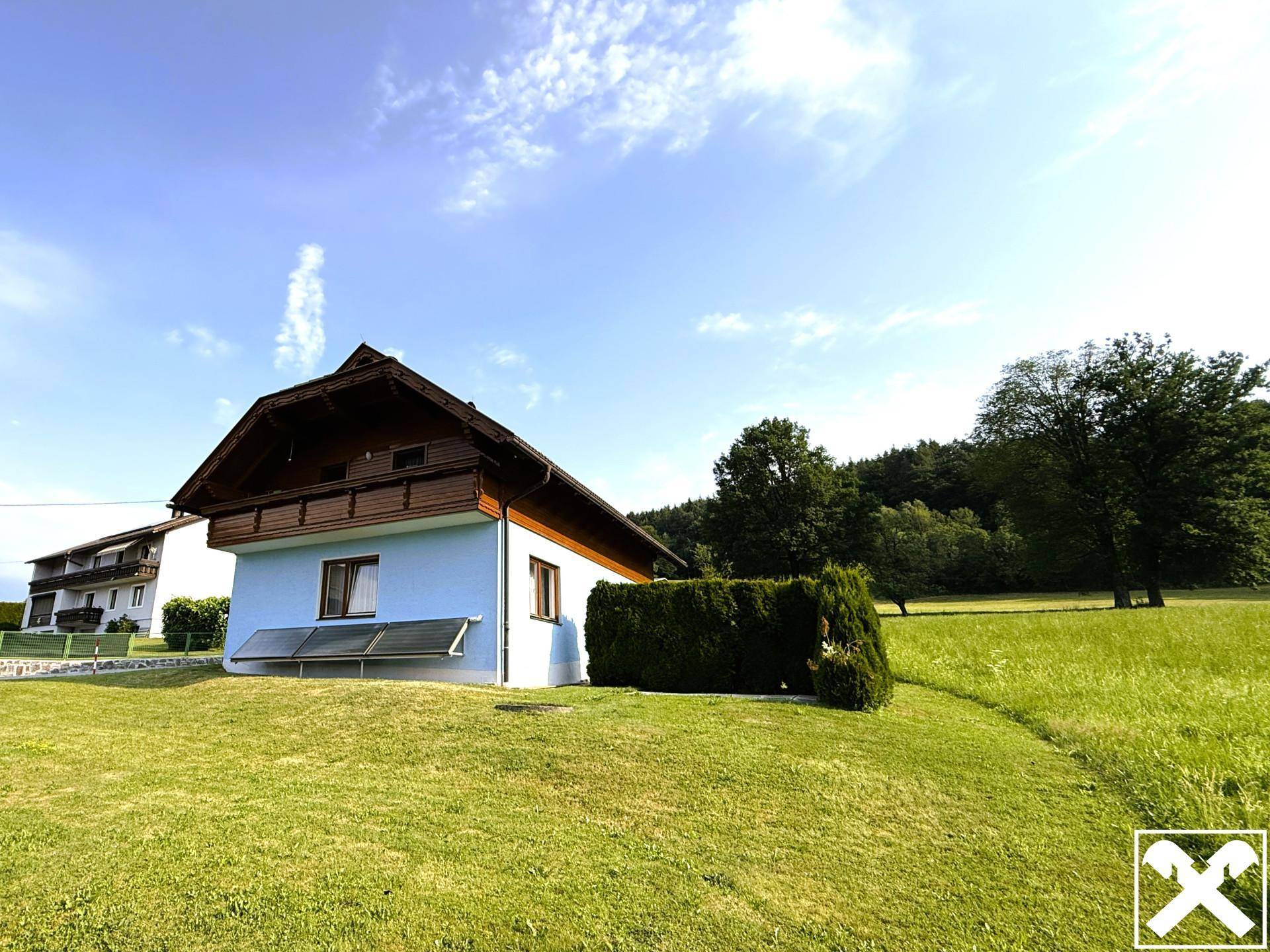 Haus in ländlicher Umgebung mit blauer Fassade, Holzverkleidung und Solarpaneelen, umgeben von Wiesen und Bäumen.