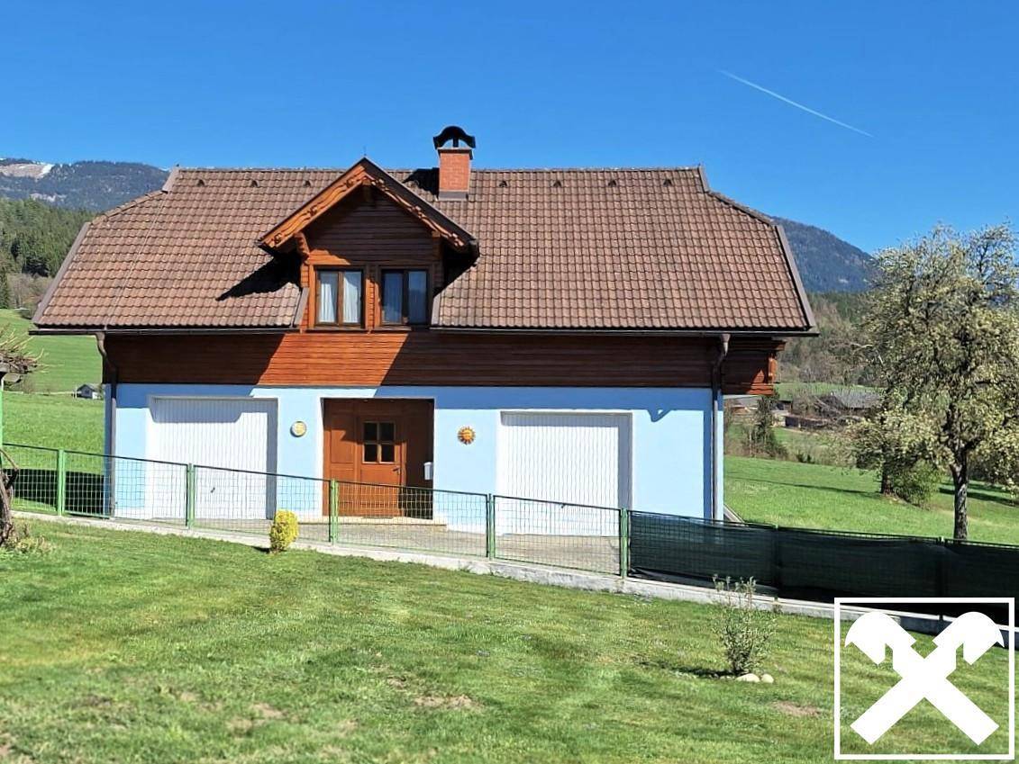 Freistehendes Haus mit blauer Fassade und Holzverkleidung, umgeben von grünen Wiesen und Bergen im Hintergrund.