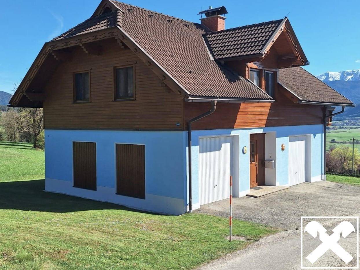 Seitenansicht des Hauses mit blauer Fassade, Holzverkleidung, Garagen und weitem Blick auf die umliegende Landschaft.