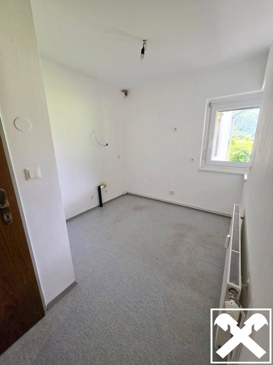 Einfaches Zimmer mit grauem Bodenbelag, weiß gestrichenen Wänden und einem Fenster.