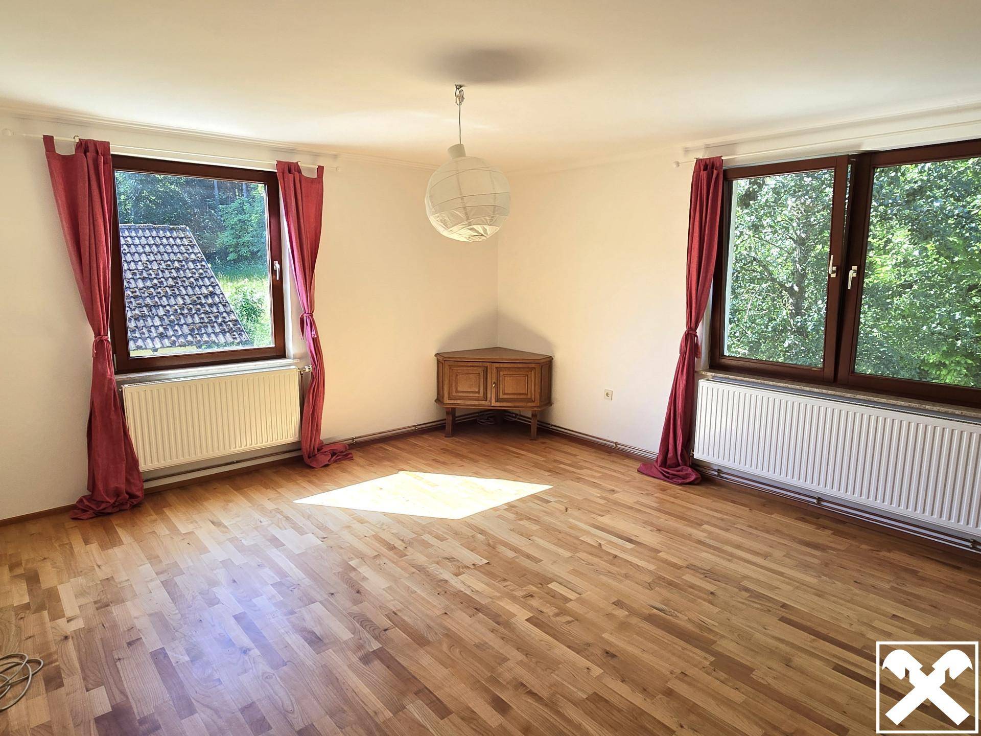 Geräumiges Zimmer mit Parkettboden, roten Vorhängen und Blick auf die bewaldete Umgebung.