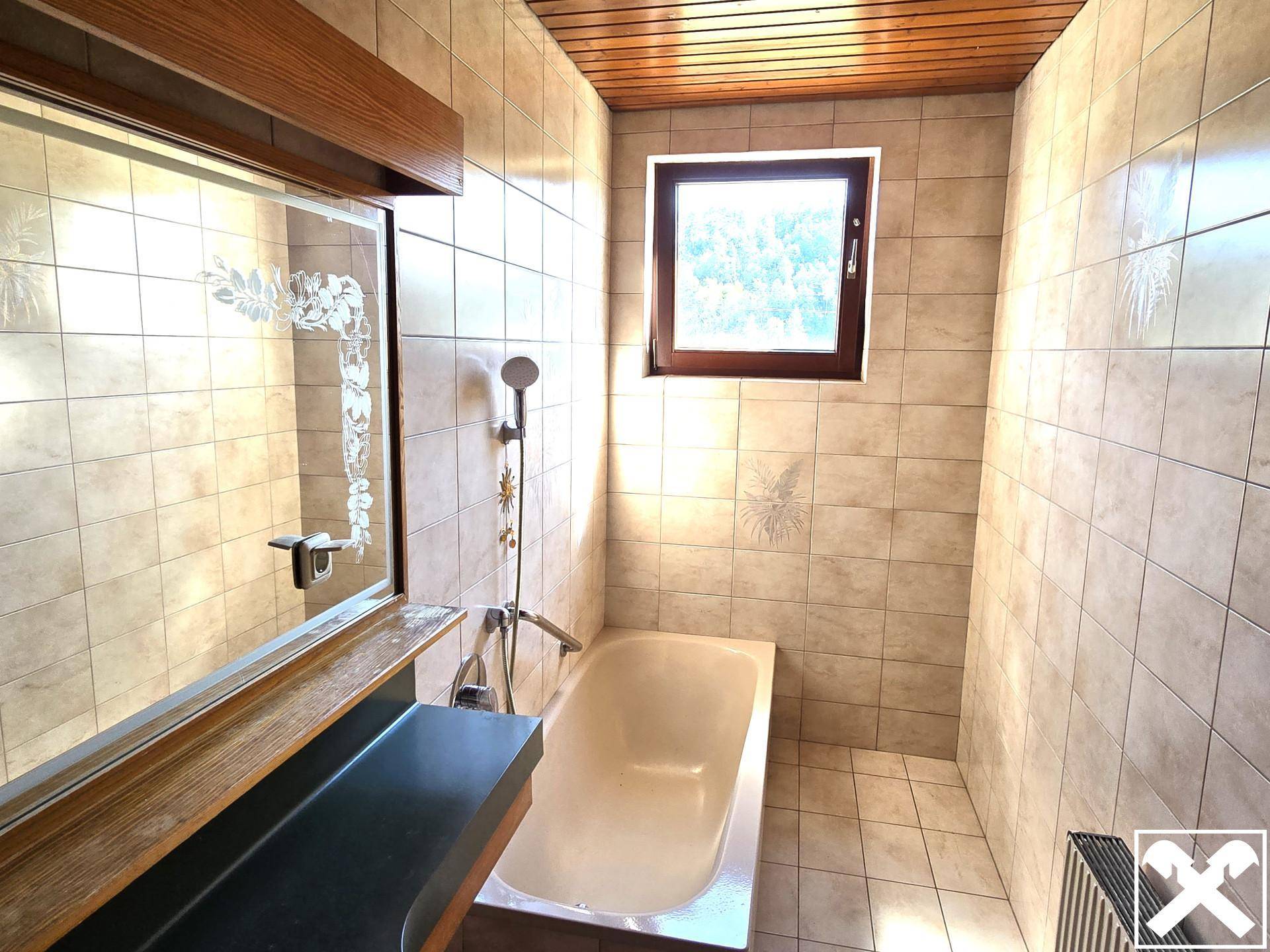 Badezimmer mit beigen Fliesen, Badewanne und Holzdecke, gut beleuchtet durch ein Fenster.