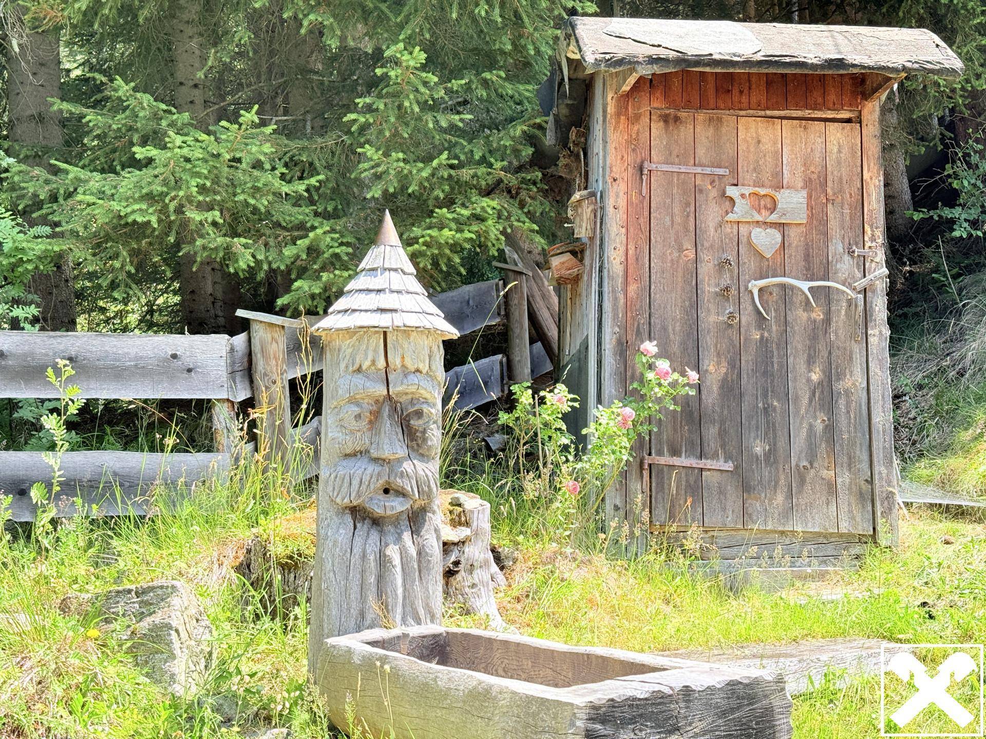 Rustikales Holzhäuschen mit Schnitzereien und einem Holzbrunnen in natürlicher Umgebung.