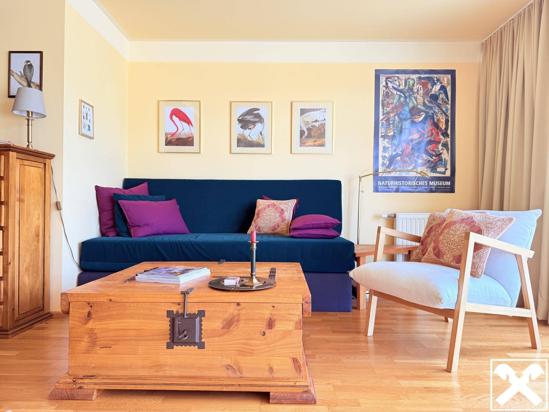 Gemütlicher Wohnbereich mit blauem Sofa, rustikalem Holztisch und farbenfrohen Bildern an der hellen Wand.