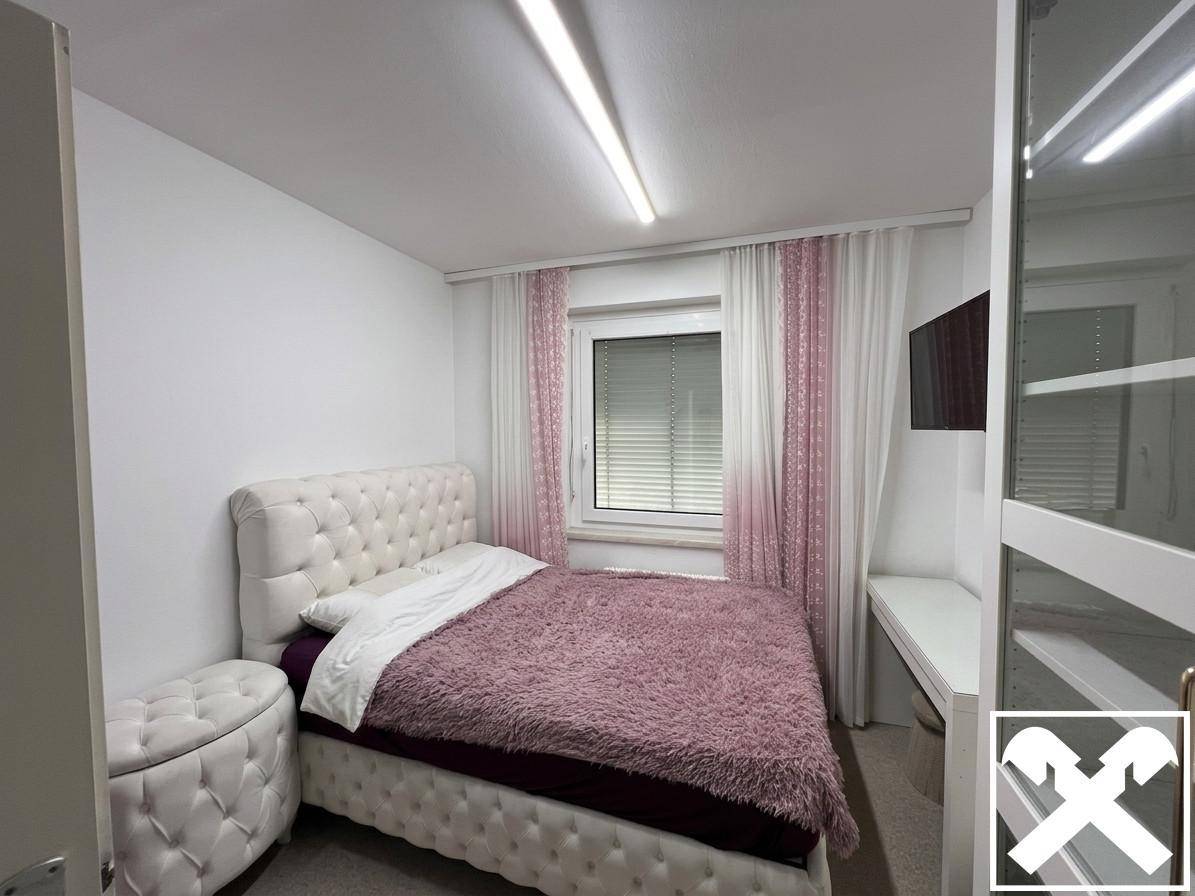 Gemütliches Schlafzimmer mit gepolstertem Bett, flauschiger Decke, Fenster und Wandfernseher.