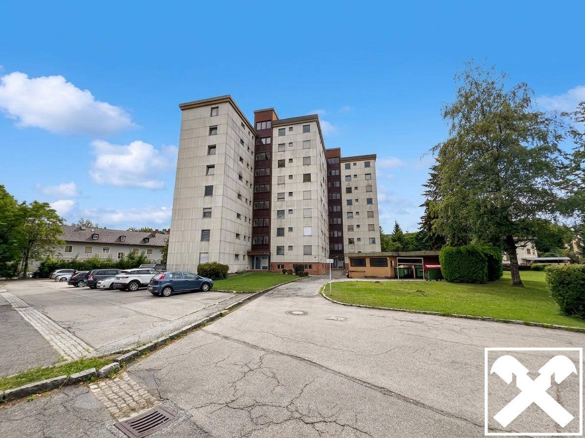 Mehrstöckiges Wohngebäude mit Parkplätzen, Bäumen und blauem Himmel an einem sonnigen Tag.
