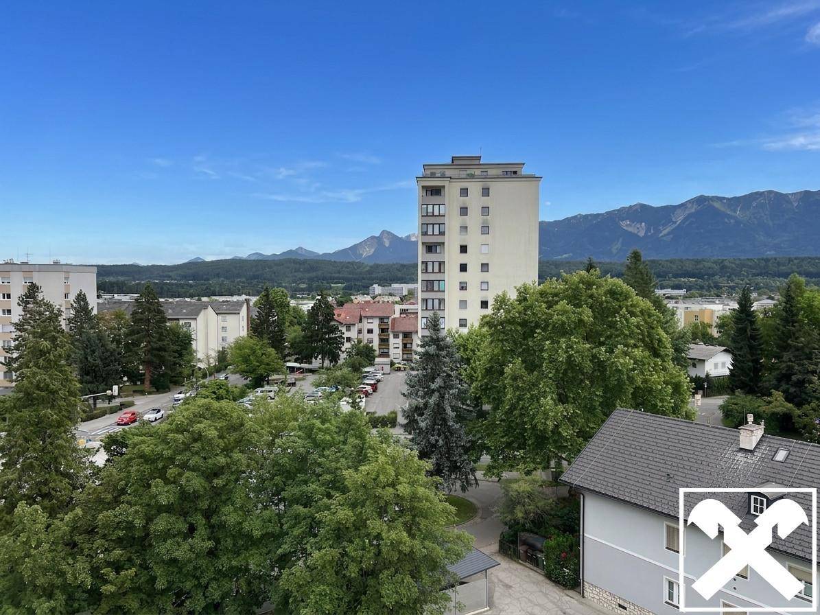 Weitblick von oben auf Wohngebäude, grüne Bäume, Parkplätze und die umliegende Berglandschaft.