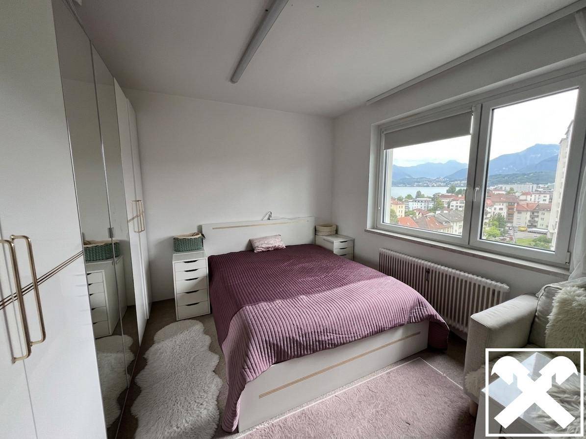 Helles Schlafzimmer mit Seeblick, lila Bettwäsche, weißen Schränken und großem Fenster.