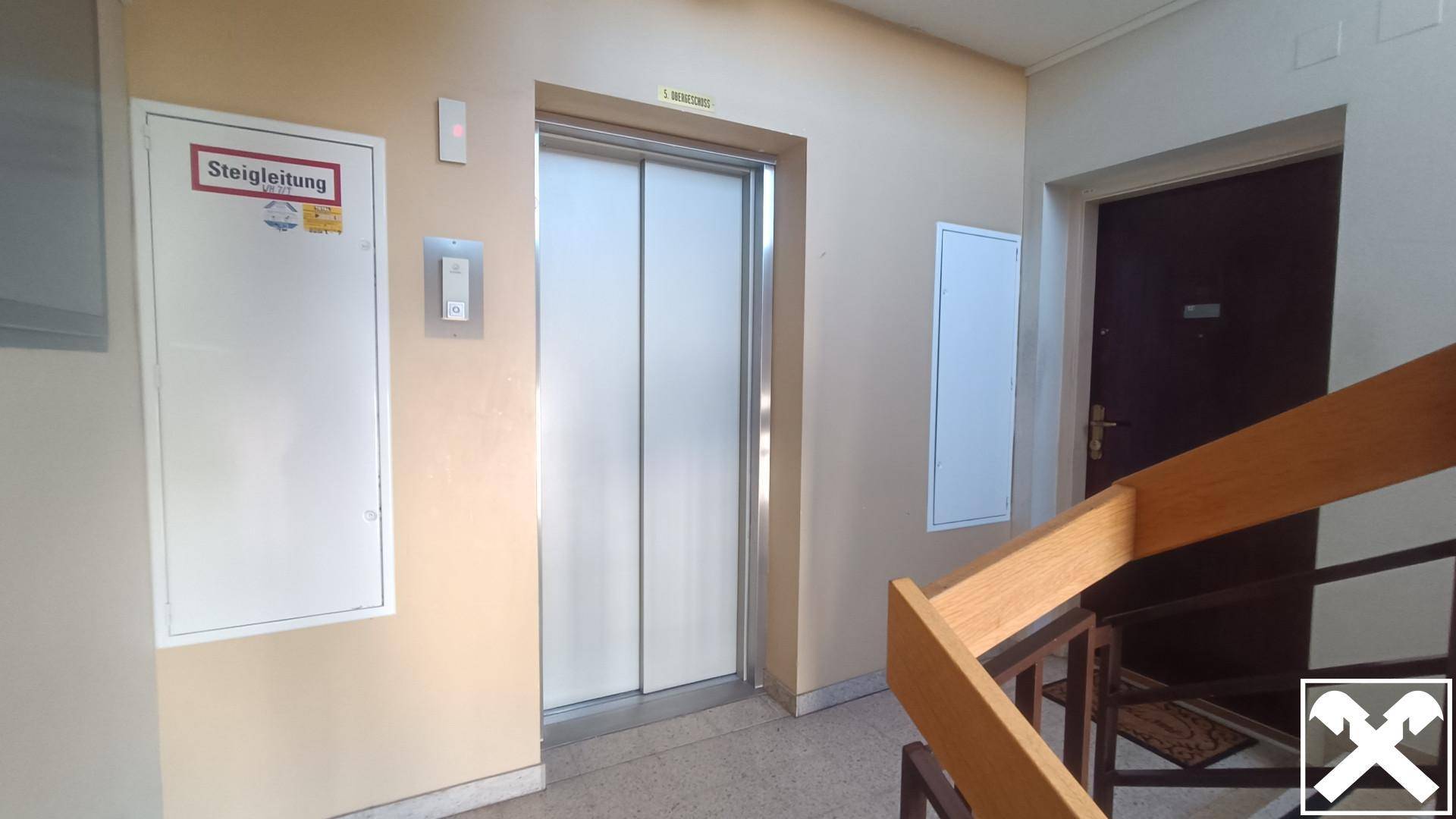 Flur im Gebäude mit Aufzug, Treppenhaus und Holzgeländer für den Zugang zu den Etagen.