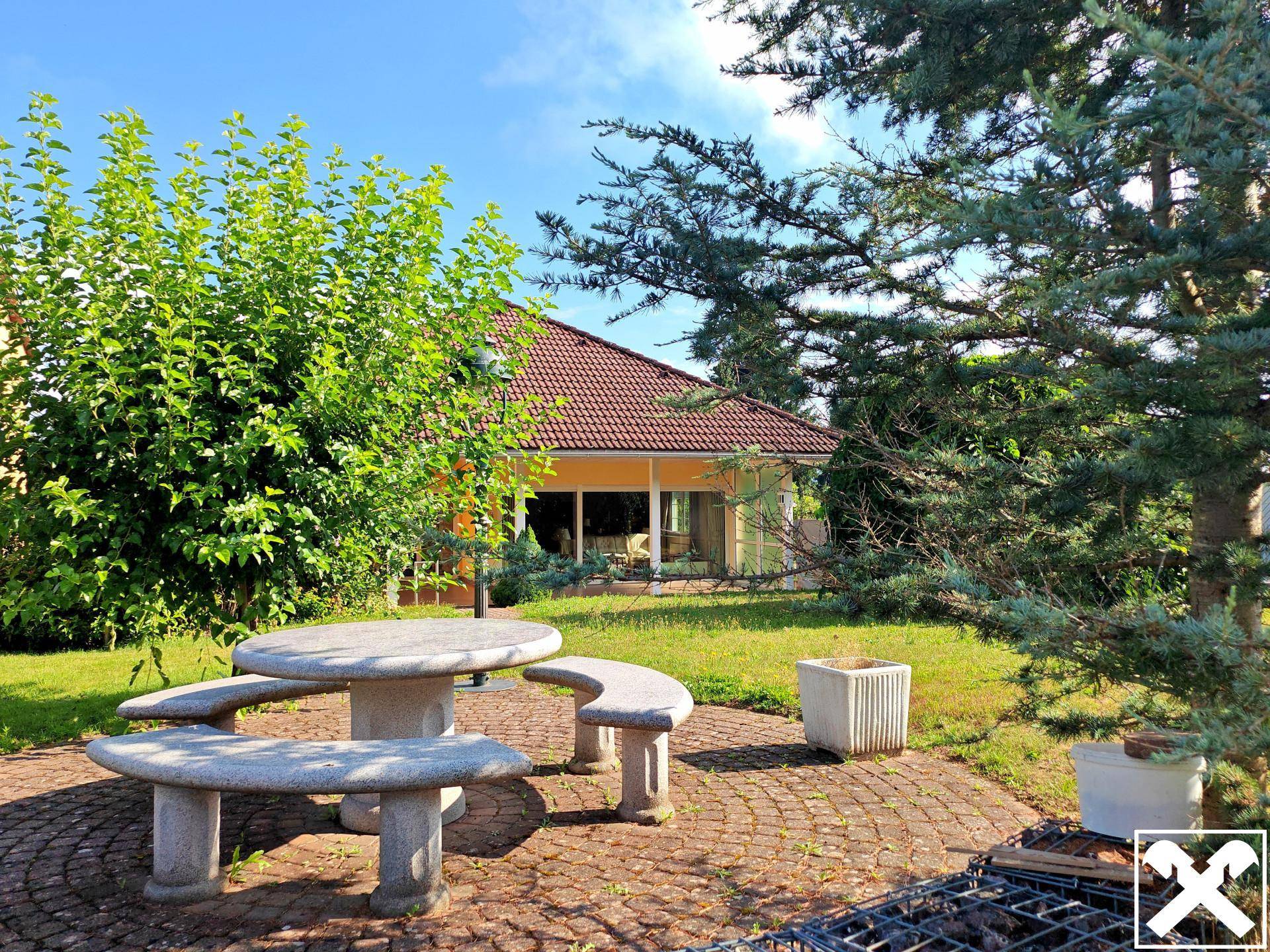 Gemütlicher Sitzbereich im Garten mit Steintisch und Grillmöglichkeit, umgeben von Bäumen.