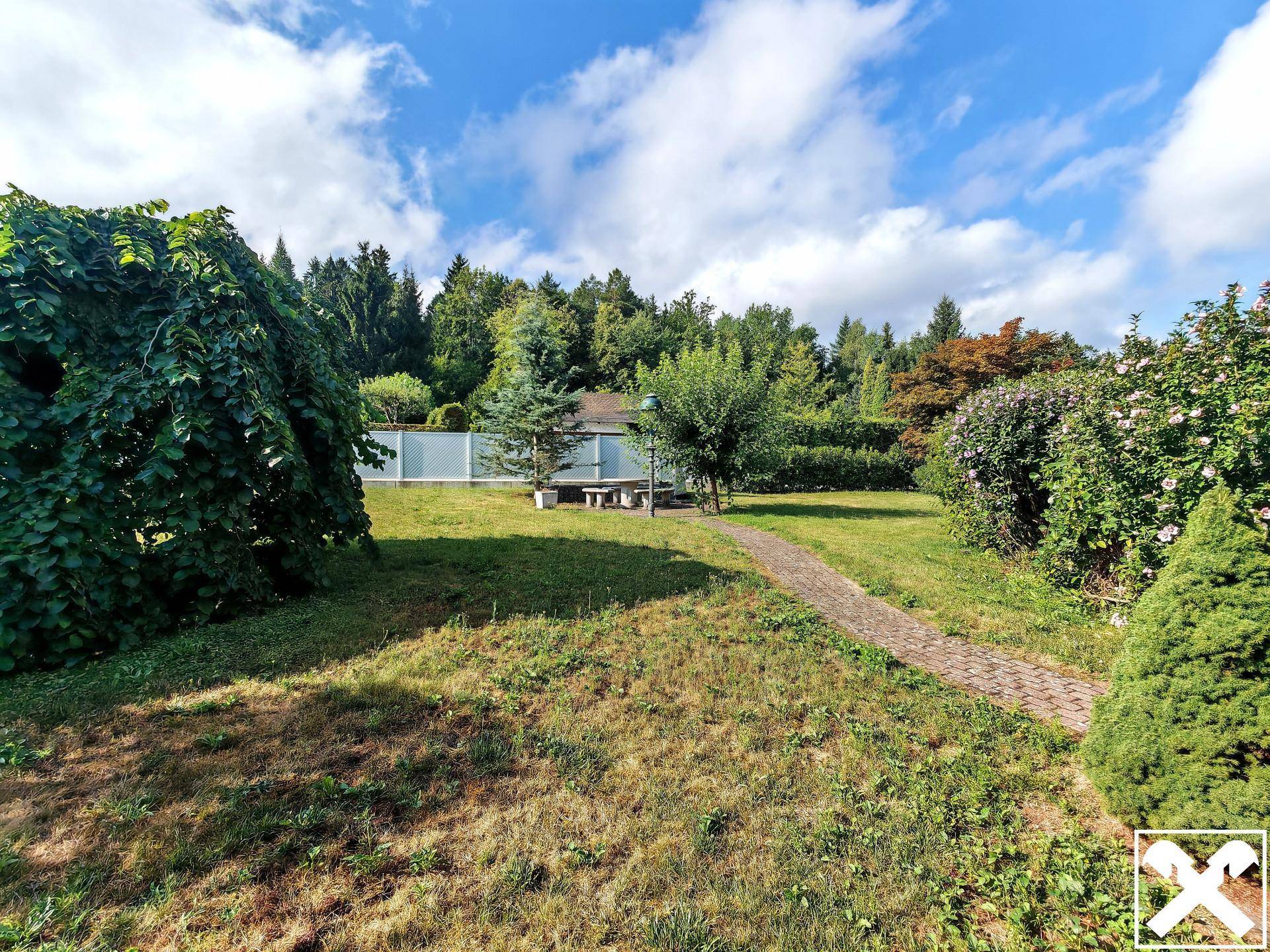 Großer Garten mit Rasenfläche, Bäumen und einem Sitzbereich im Hintergrund unter blauem Himmel.