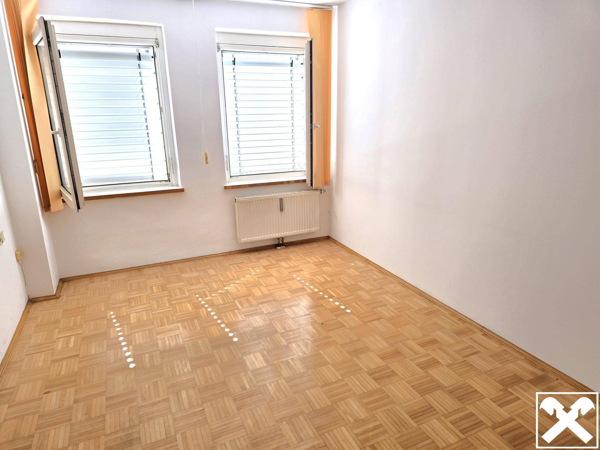 Heller, unmöblierter Raum mit Parkettboden, zwei Fenstern und Heizkörper.