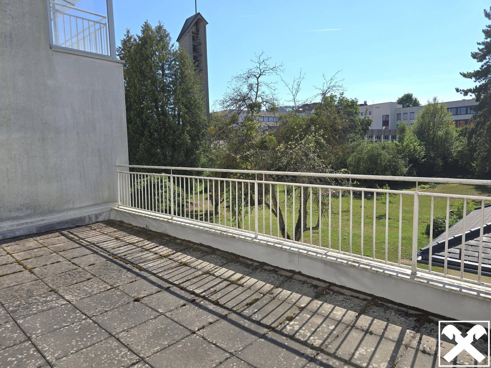 Große geflieste Terrasse mit Metallgeländer und Blick ins Grüne sowie auf einen Kirchturm.