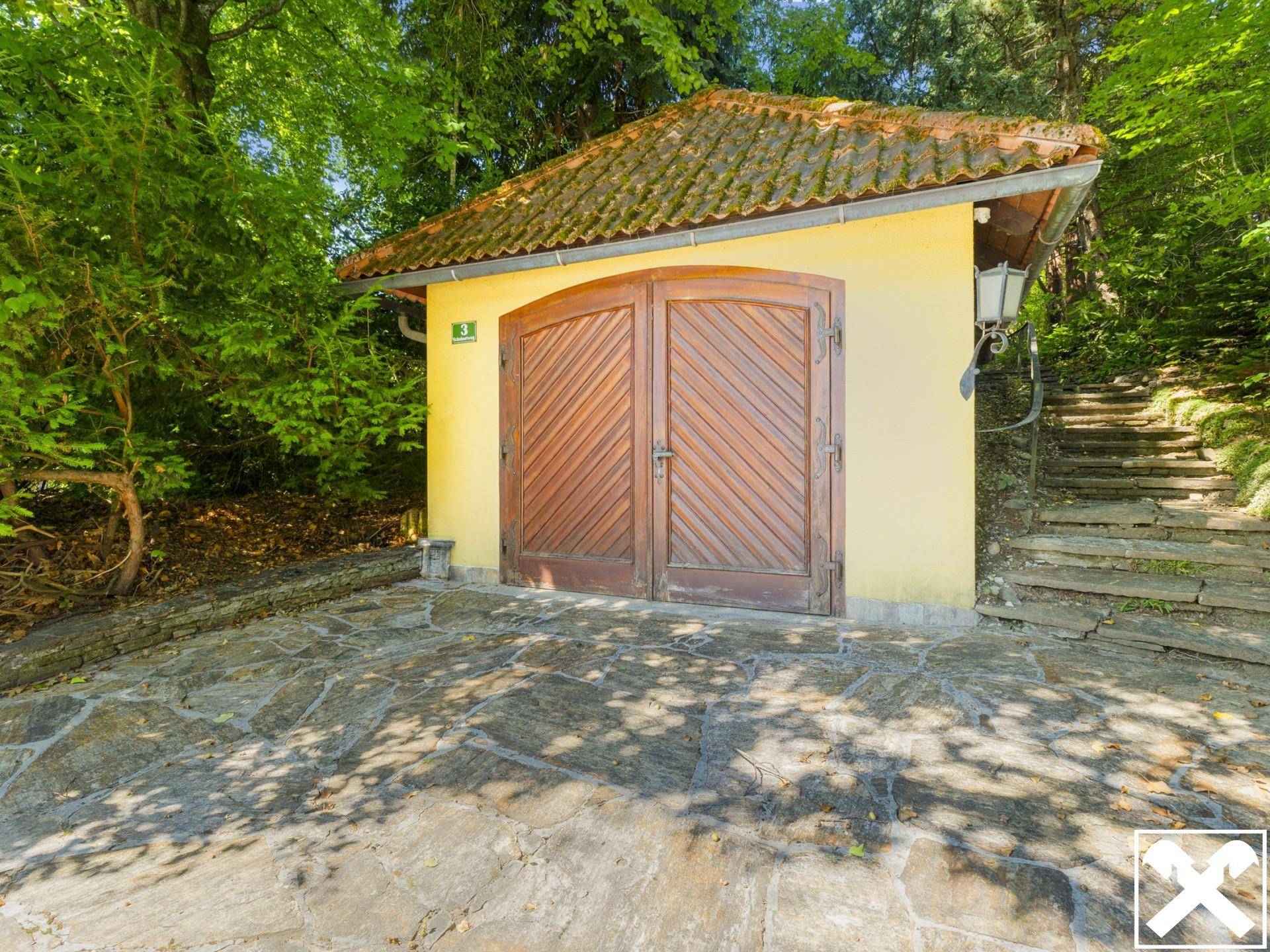 Separate Garage mit Holztoren und Natursteinpflasterung, eingebettet in die grüne Umgebung.