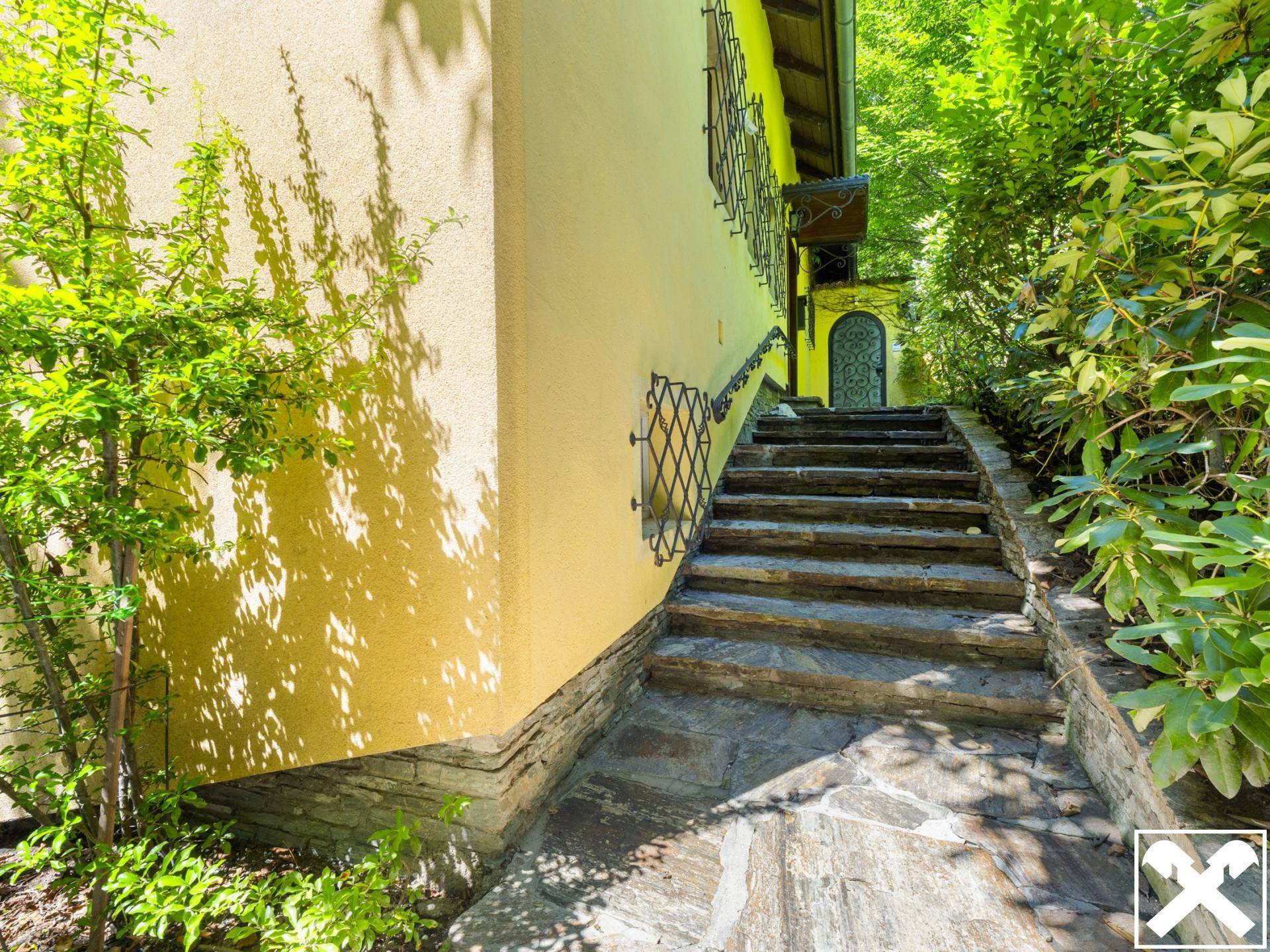 Natursteintreppe führt entlang der Hauswand hinauf, umgeben von üppigem Grün.