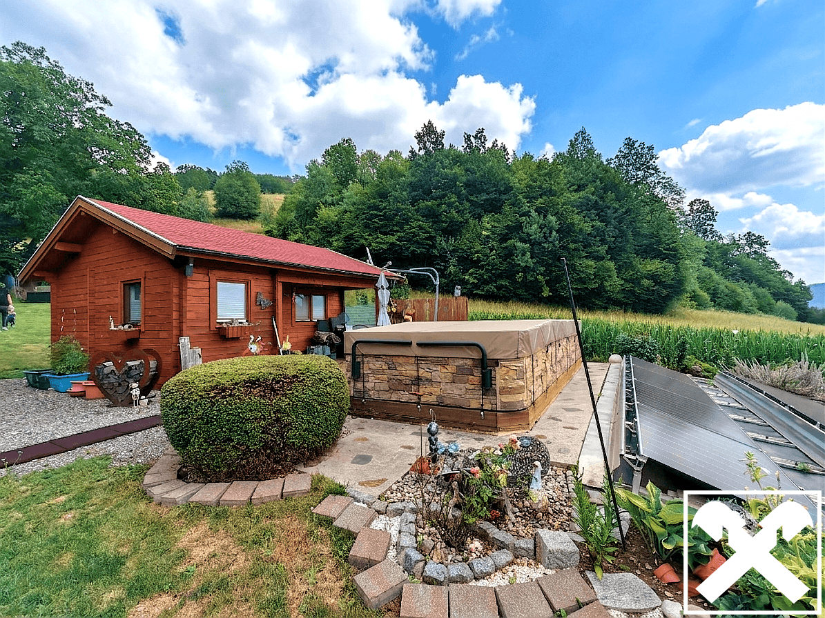 Außenansicht des Holzhauses mit überdachtem Whirlpool, Solaranlage und gepflegtem Gartenbereich vor Waldkulisse.