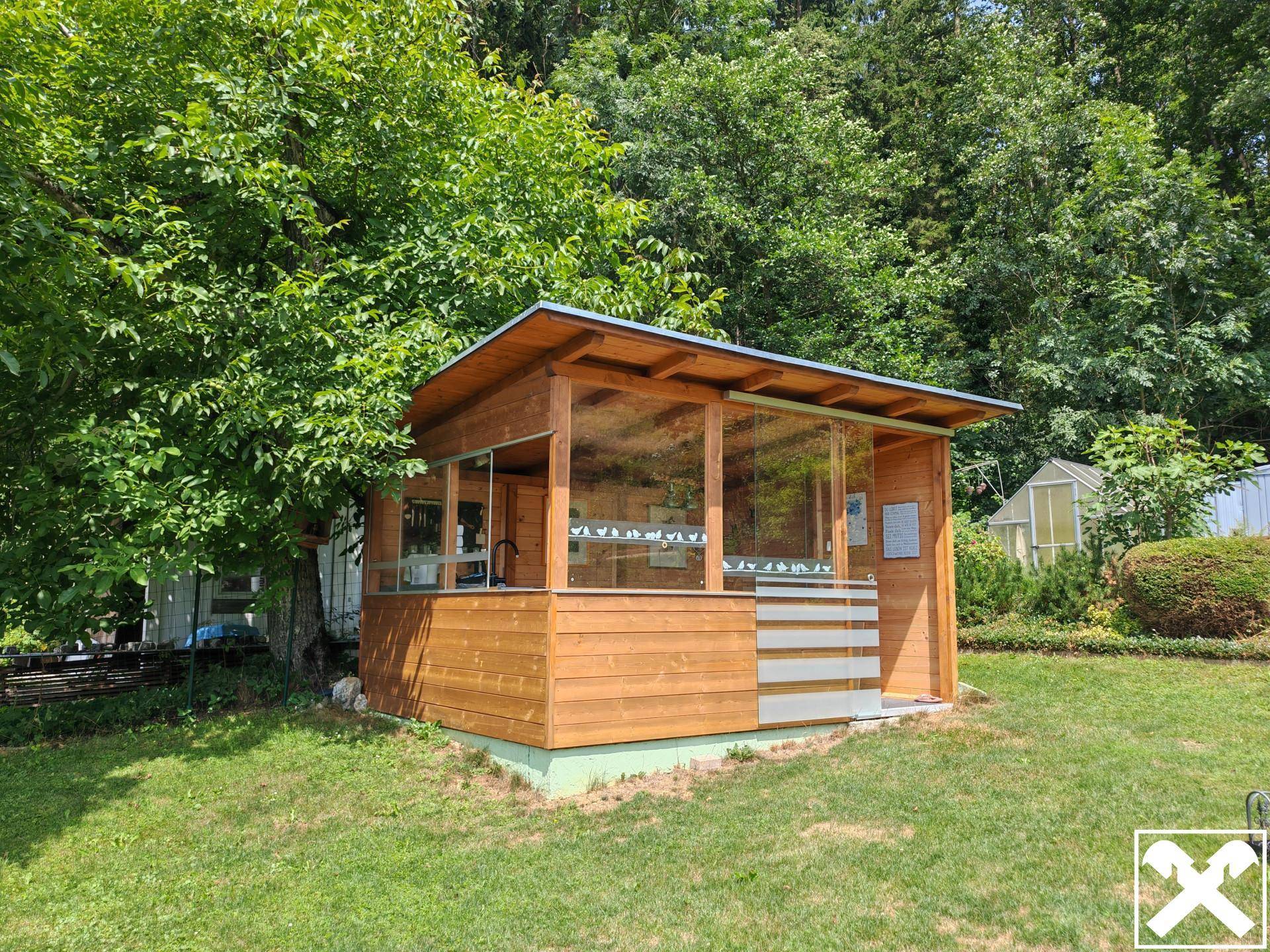Charmantes Gartenhaus aus Holz mit Glasfronten, umgeben von üppigem Grün und Rasenfläche.