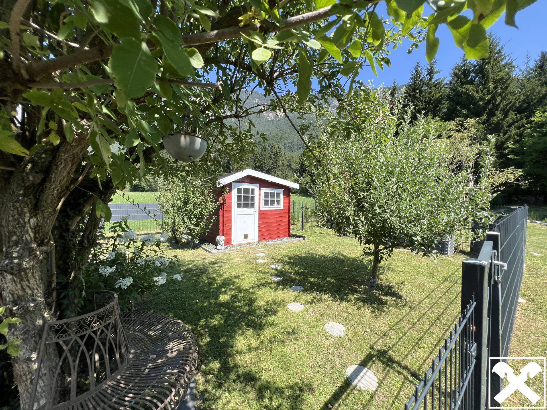 Idyllischer Garten mit kleinem roten Gartenhaus, schattenspendenden Bäumen und Bergblick.