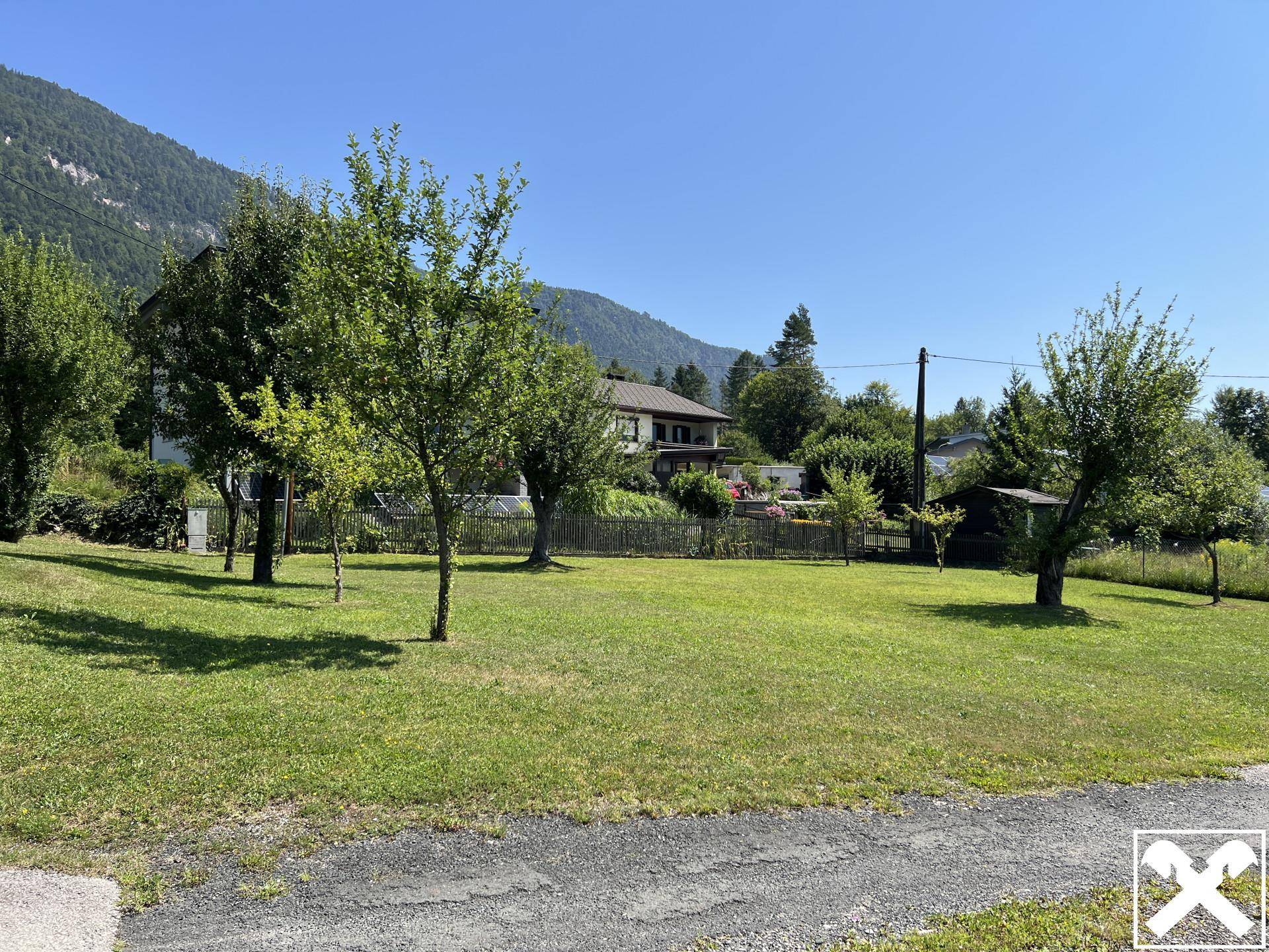 Weitläufige Rasenfläche mit jungen Bäumen und einem Wohnhaus im Hintergrund, umgeben von Natur.