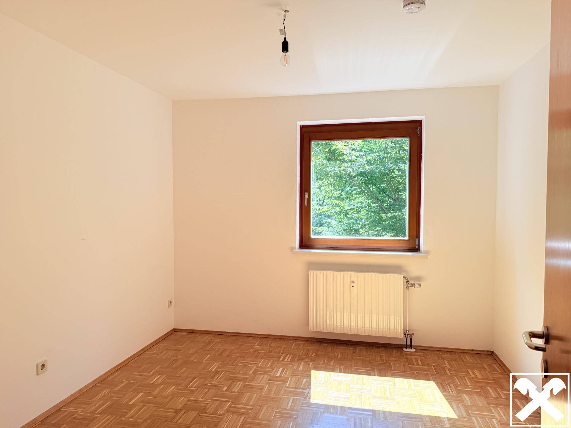 Helles Schlafzimmer mit Parkettboden, weiß gestrichenen Wänden und Fensterblick ins Grüne.
