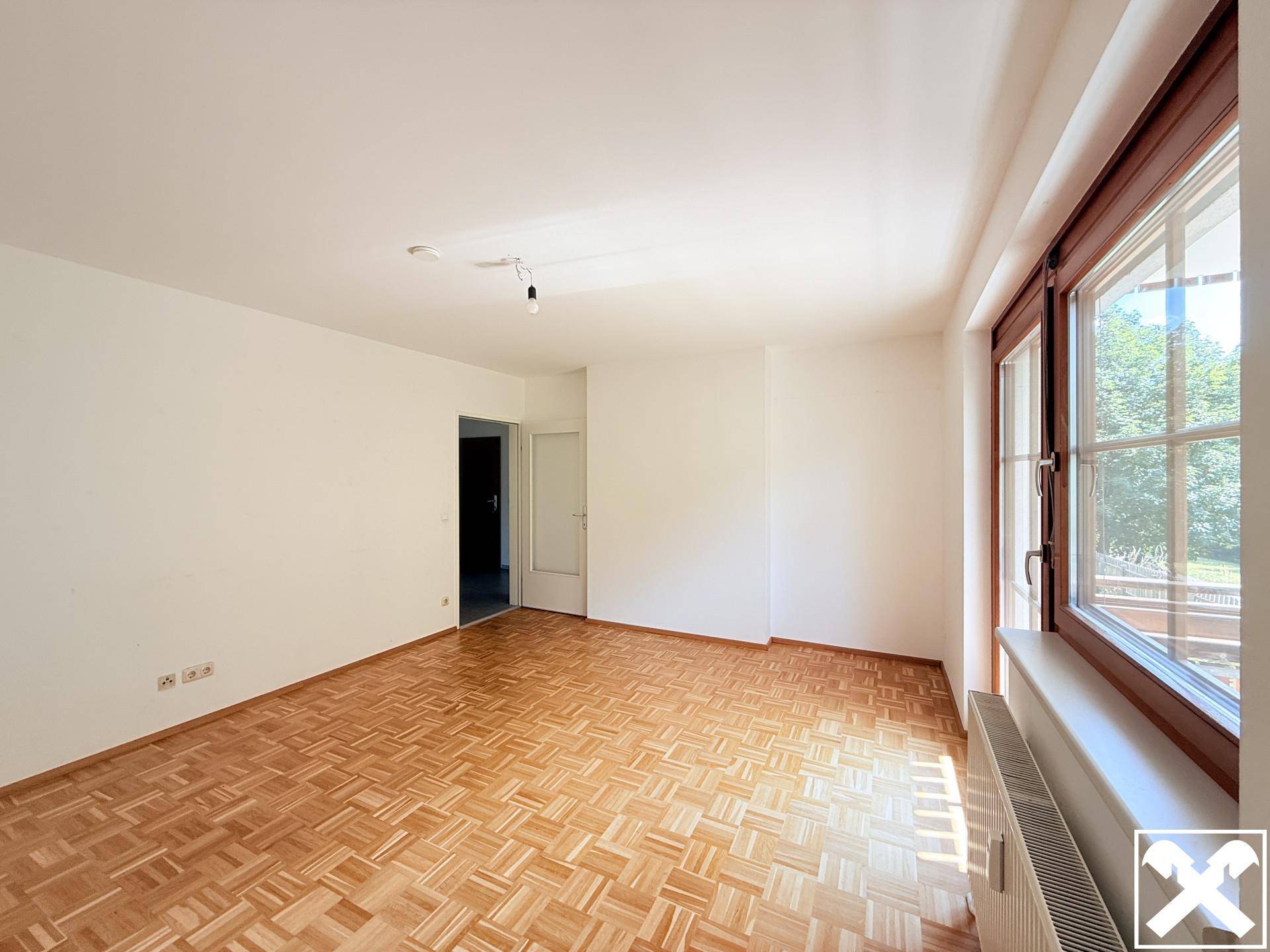 Geräumiger, leerer Wohnraum mit Parkettboden und großem Fenster, das viel Tageslicht hereinlässt.