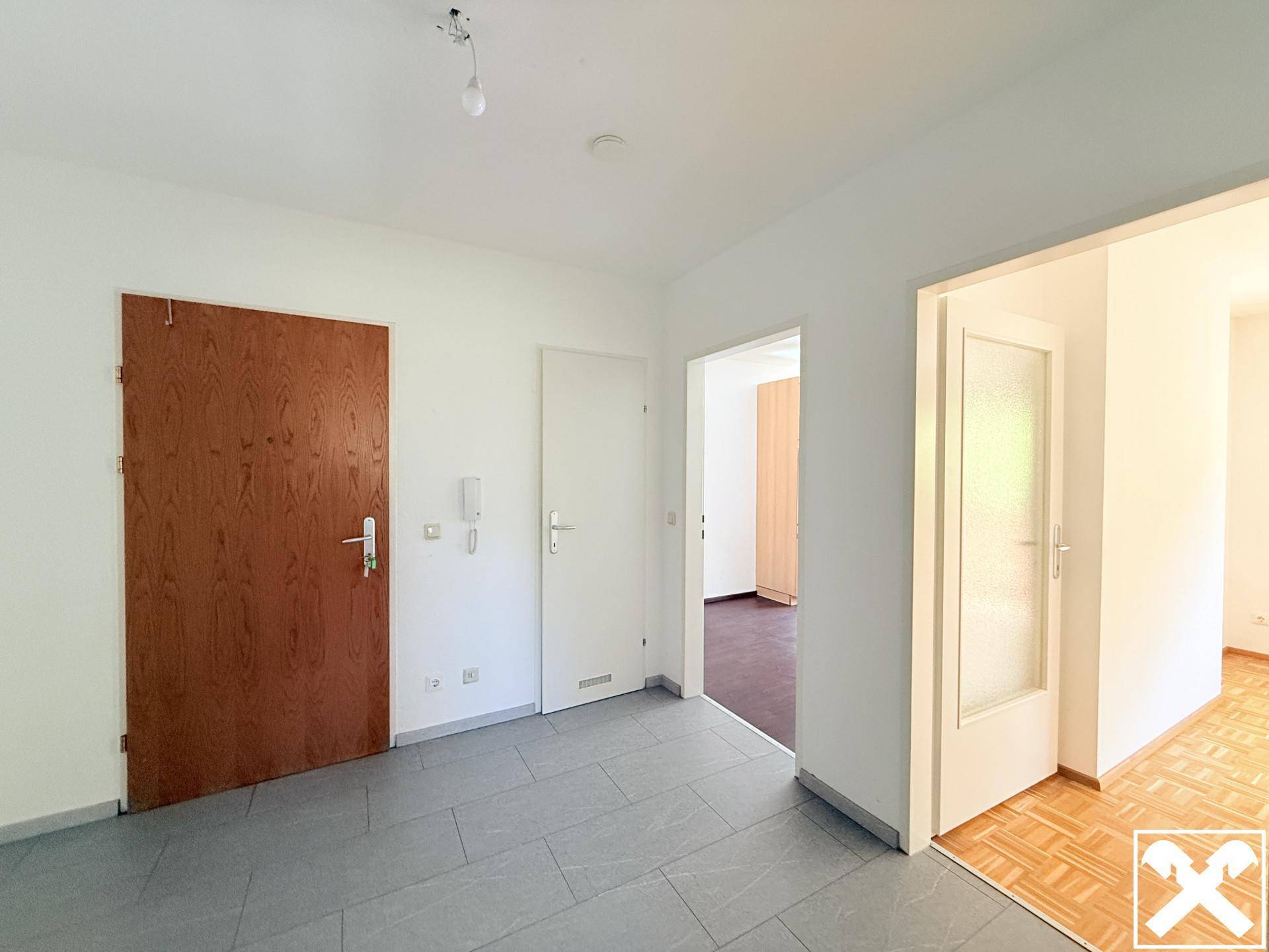 Eingangsbereich mit grauen Fliesen, einer Holztür und Zugang zu weiteren Räumen.