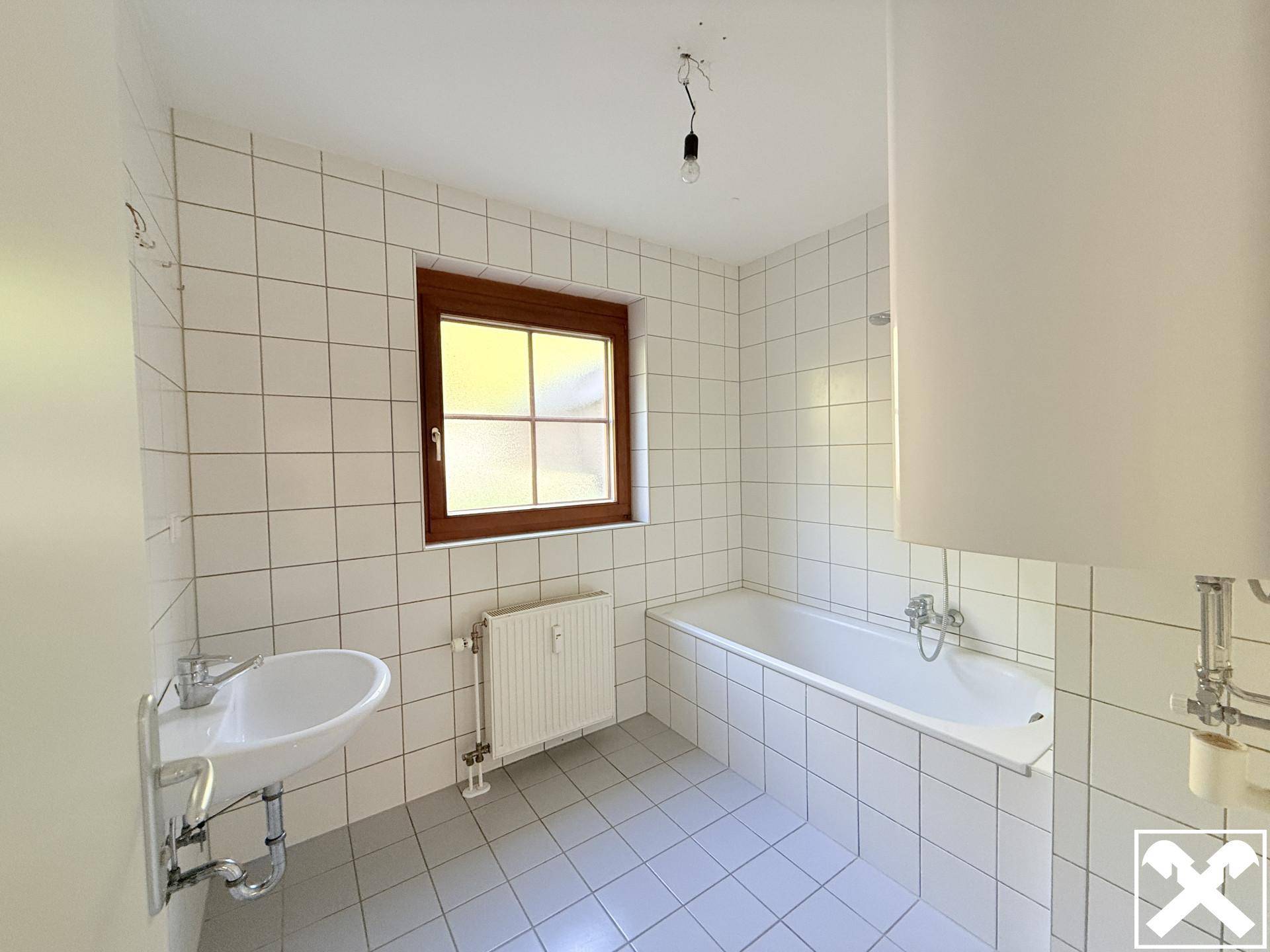 Badezimmer mit weißen Fliesen, Badewanne, Waschbecken und einem Fenster für Tageslicht.