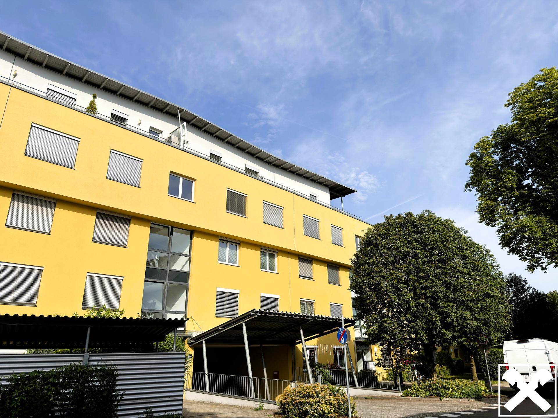 Modernes gelbes Gebäude mit Flachdach und Bäumen unter blauem Himmel.