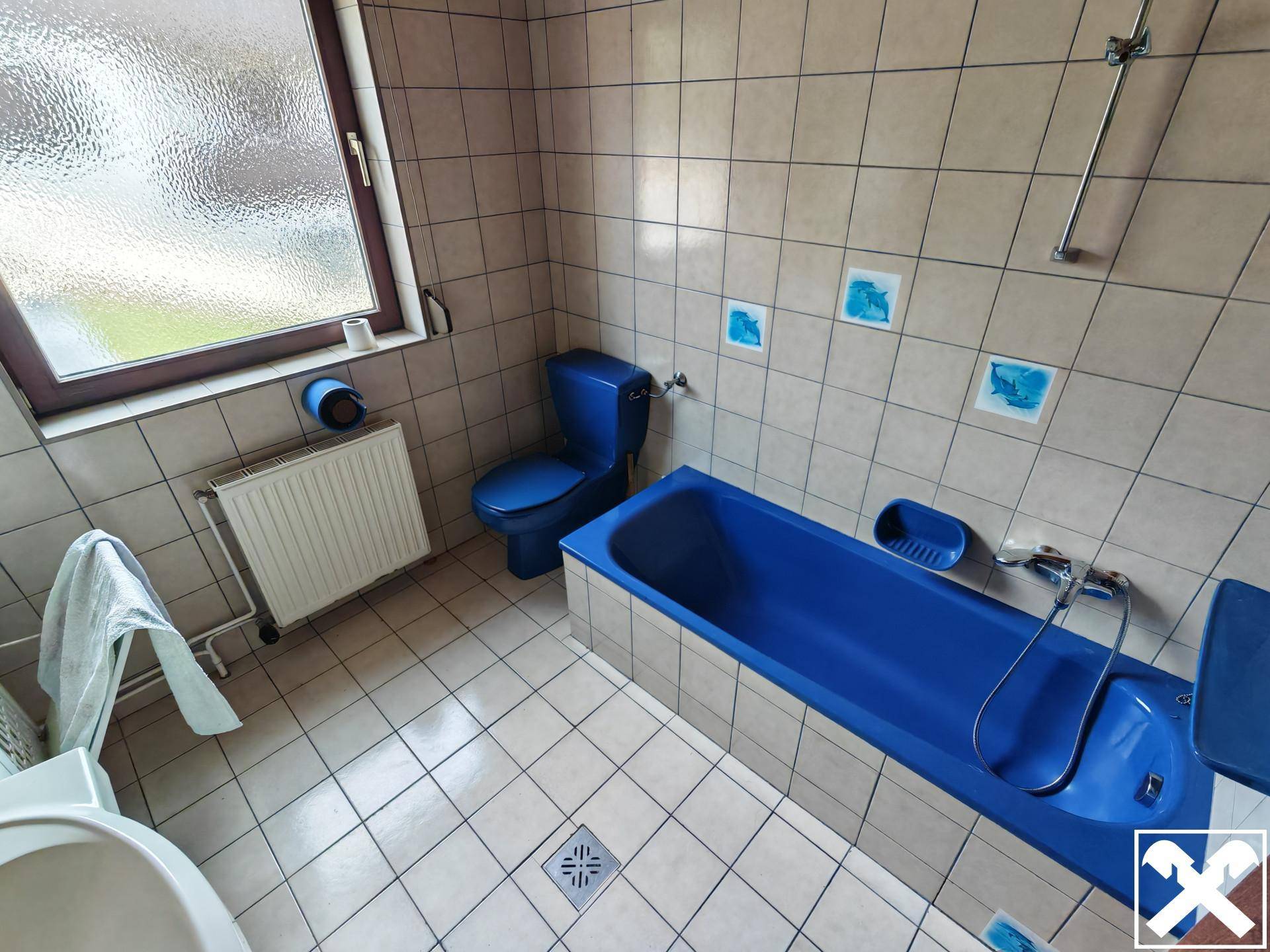 Badezimmer mit blauer Badewanne und WC, gefliest in hellen Farben mit Fenster.