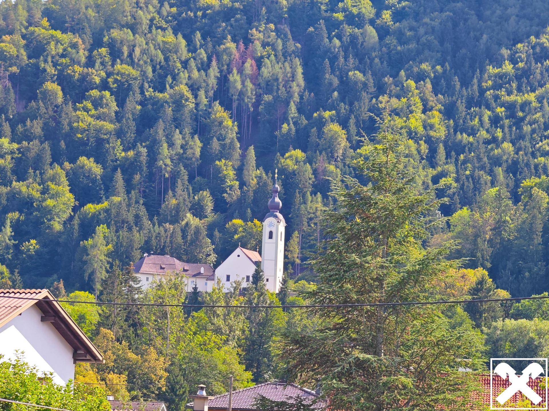Idyllischer Blick auf eine Kirche im Tal, umgeben von dicht bewaldeten Bergen und Bäumen.
