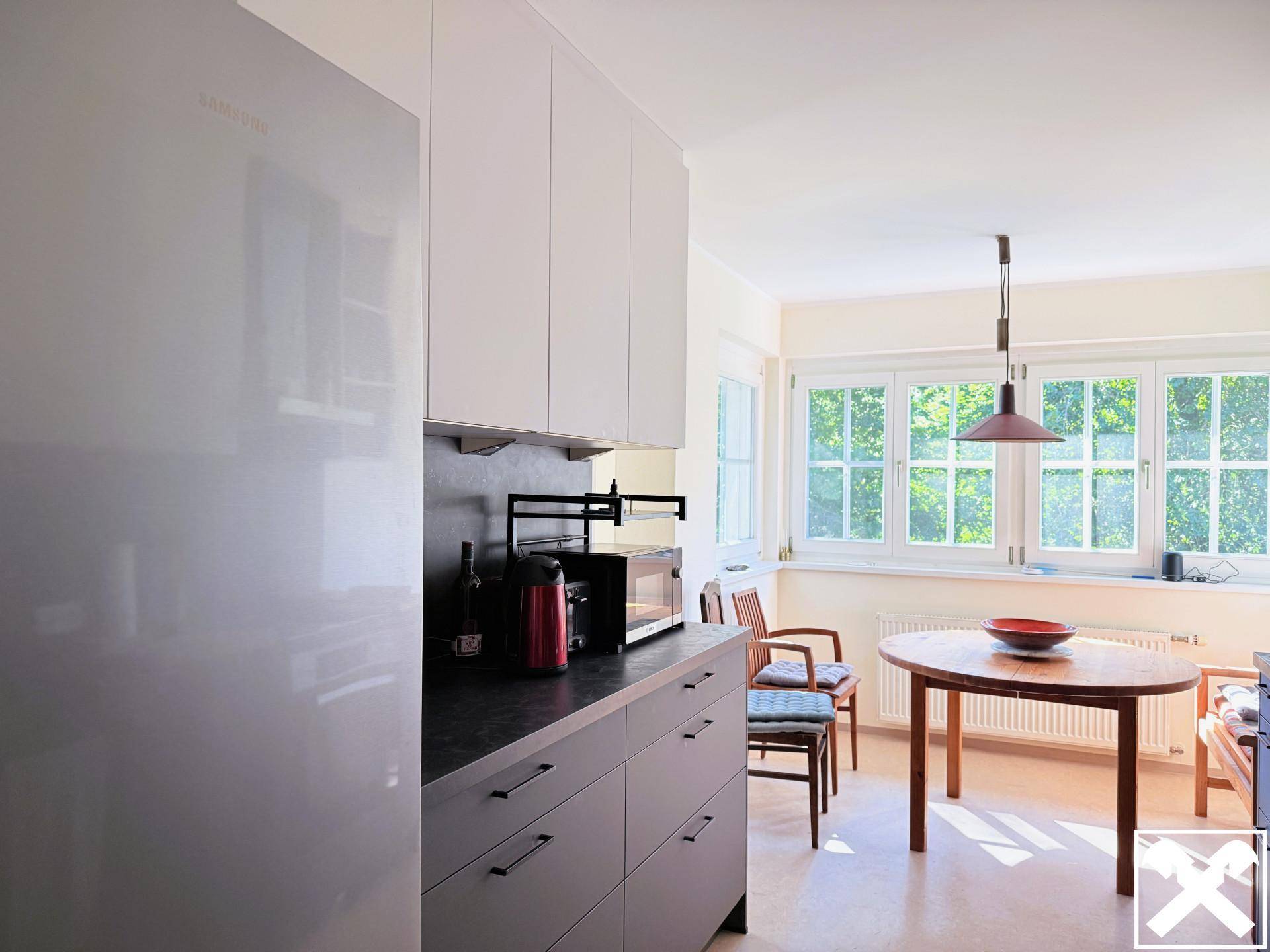 Moderne Küche mit weißen Oberschränken, dunkler Arbeitsplatte und Essbereich am Fenster.
